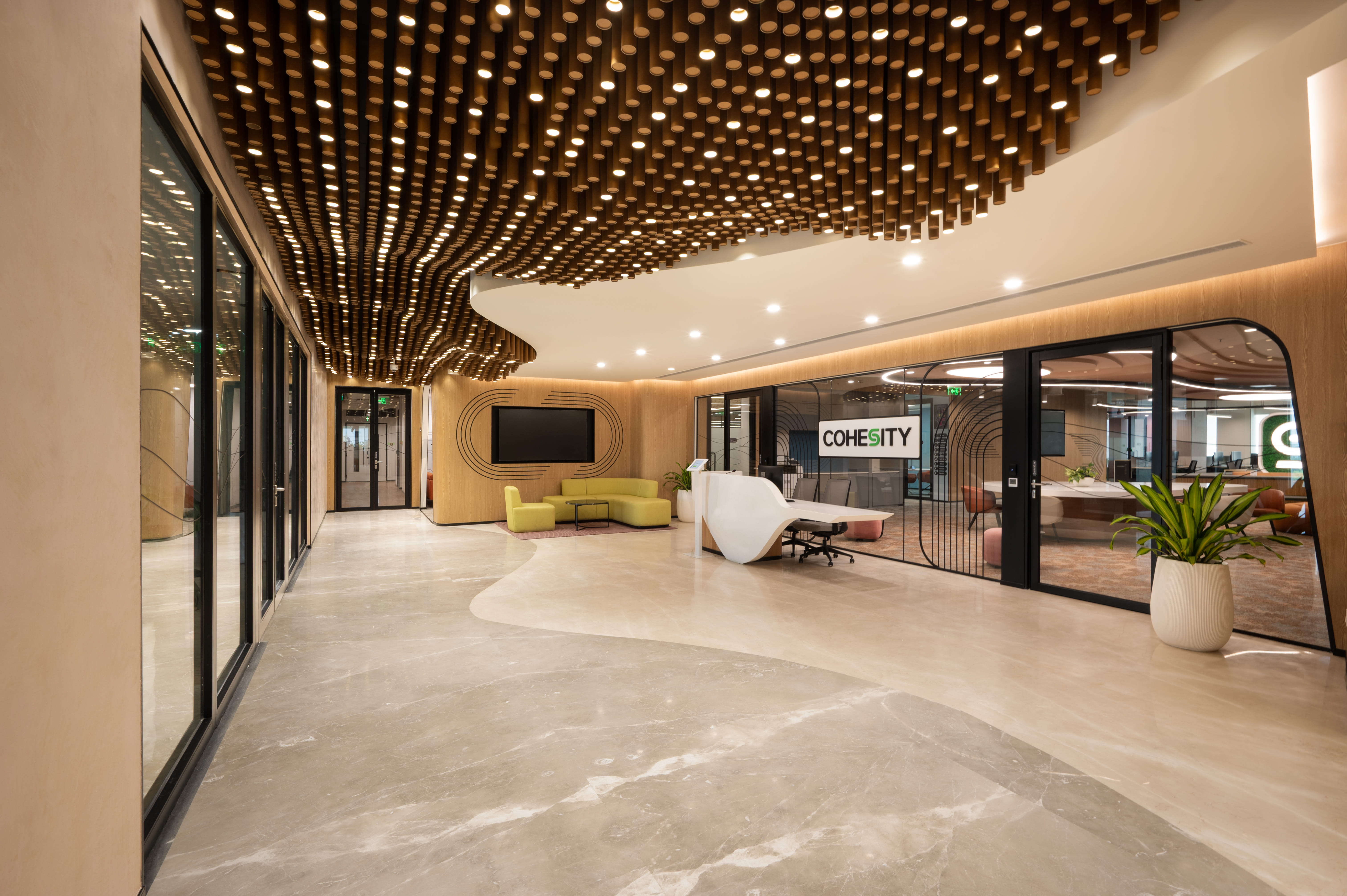 Modern office lounge area with round tables, brown chairs, a kitchenette against a brick wall, hanging lights, and a glass-walled meeting room with urban mural.