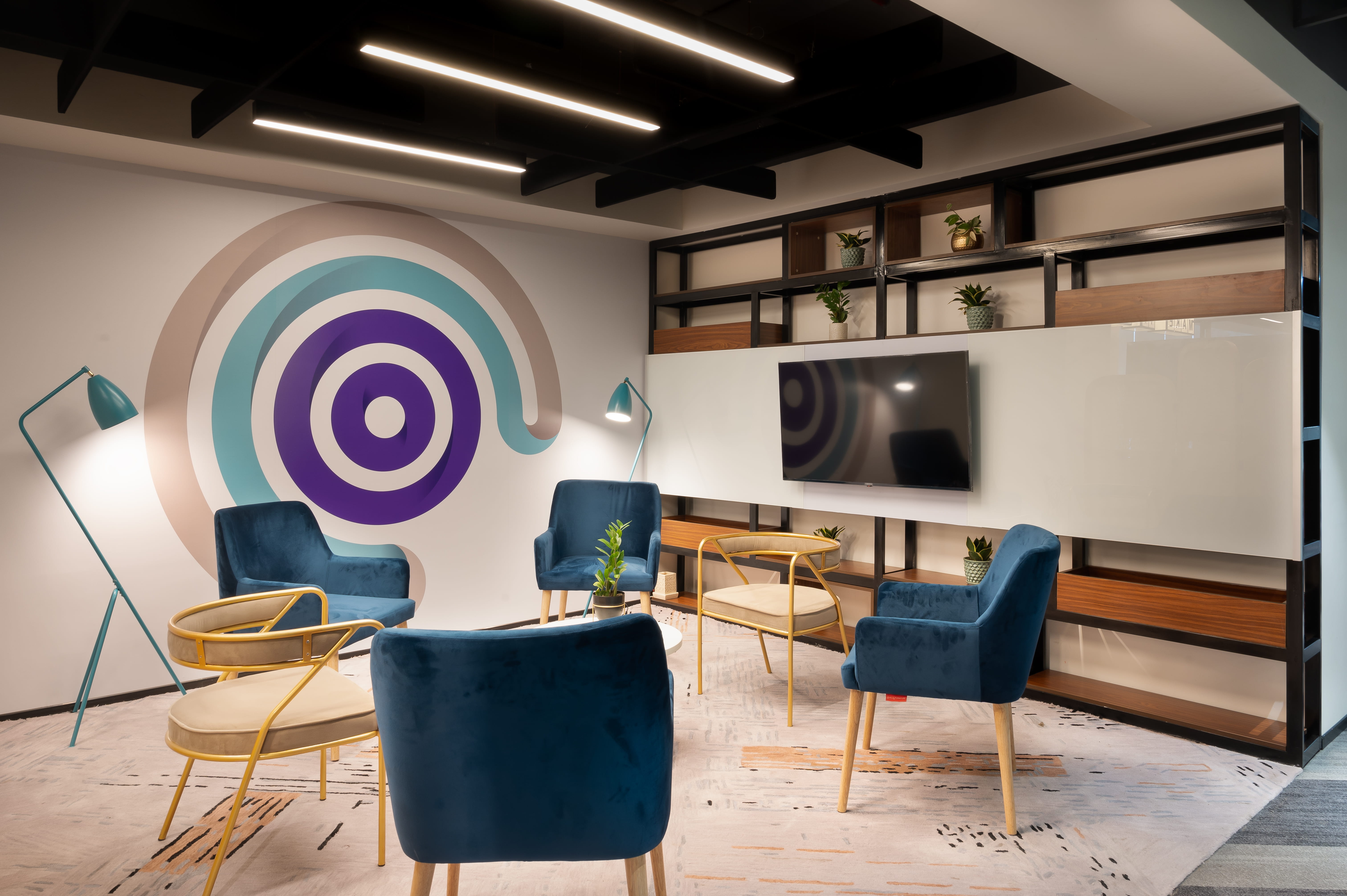 Modern café lounge area with a gray sectional sofa, red and black pillows, geometric patterned rug, two black wireframe tables, a potted plant, and a neon sign reading 'Work Café' with a coffee cup icon on a brick wall.