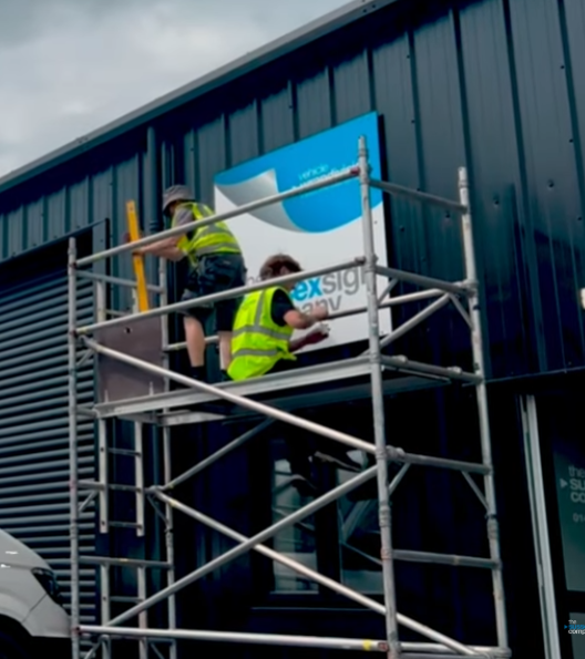 Two workers in safety vests on scaffolding install a sign on a building's exterior wall.