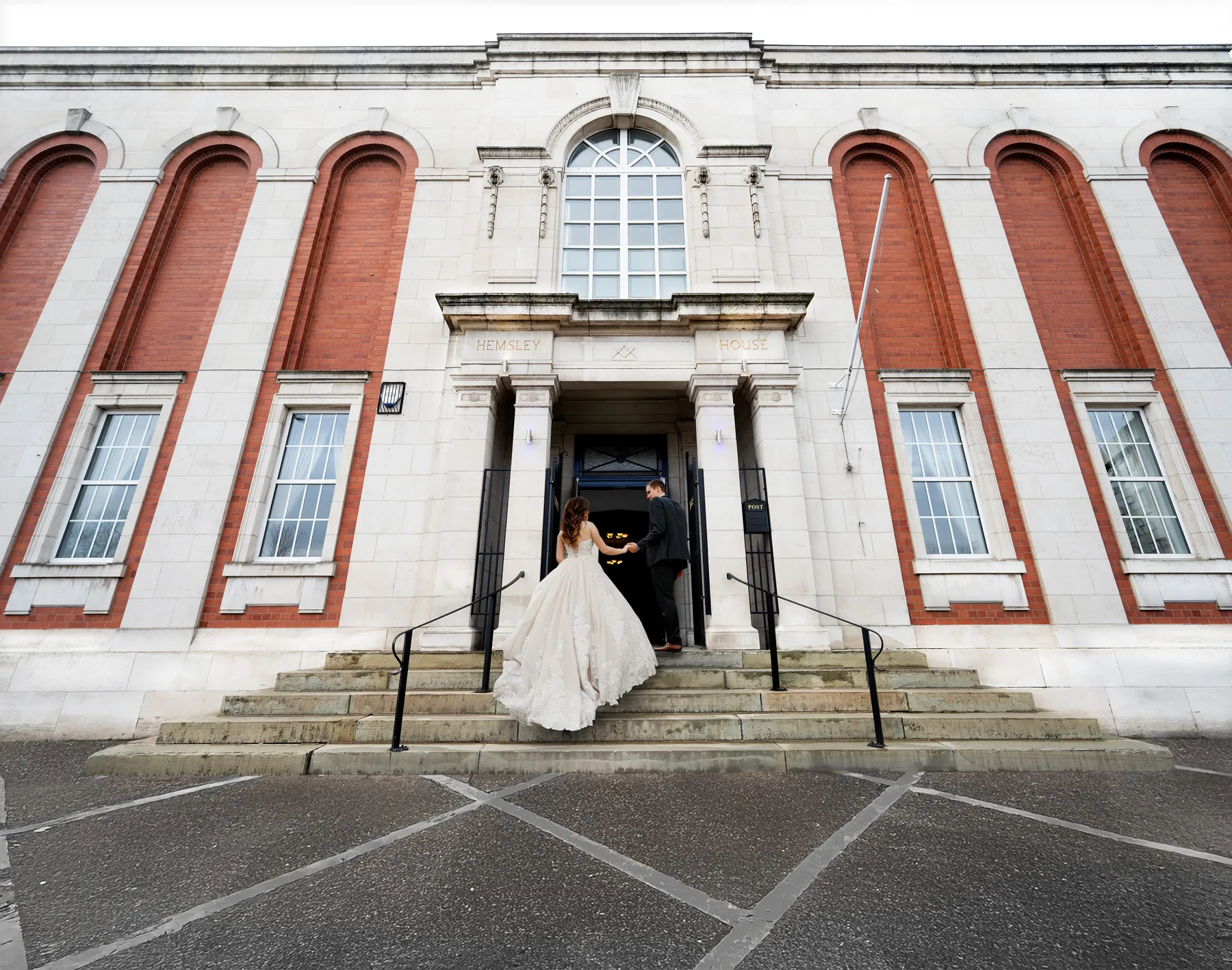 Bride in white gown and groom holding hands walking into the Hemsley House entrance.