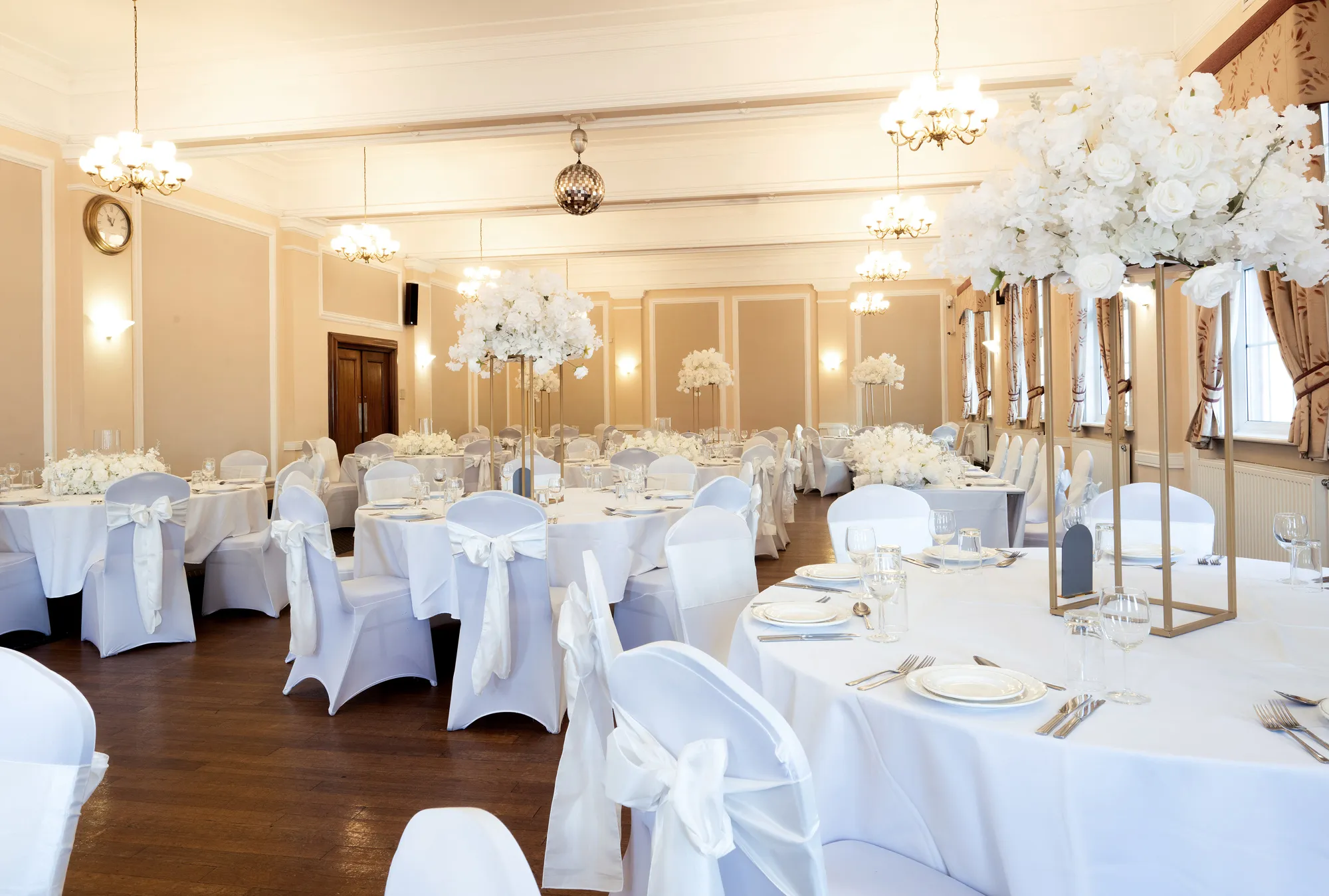 Elegant banquet hall with round tables, white chairs, and tall floral centerpieces in white and gold.