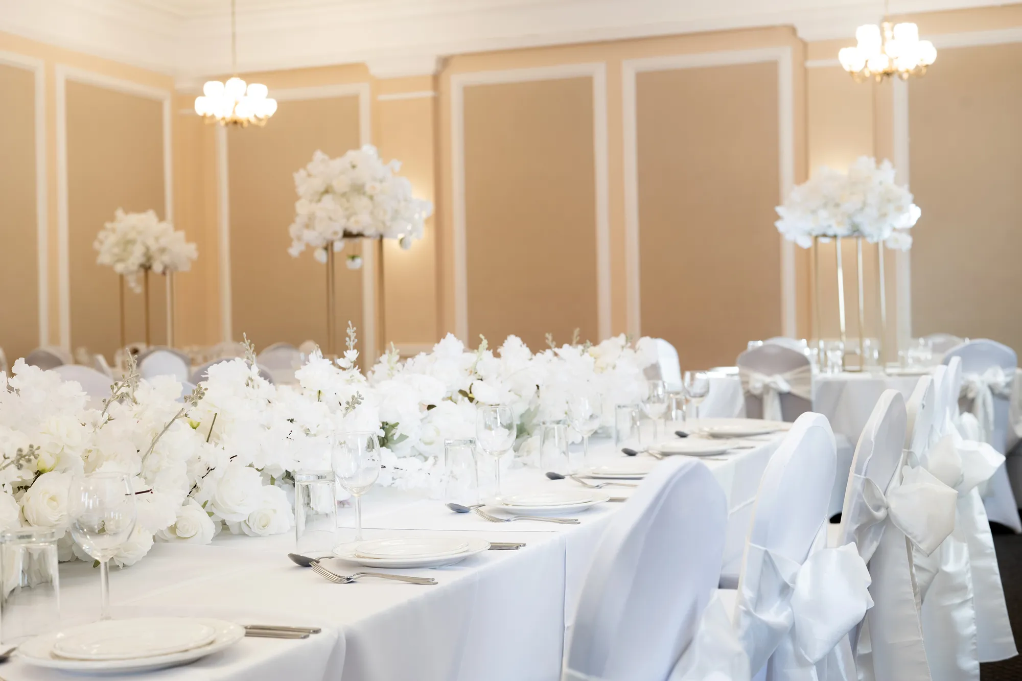 Elegant white banquet table with floral centerpieces and chairs adorned with white satin bows.