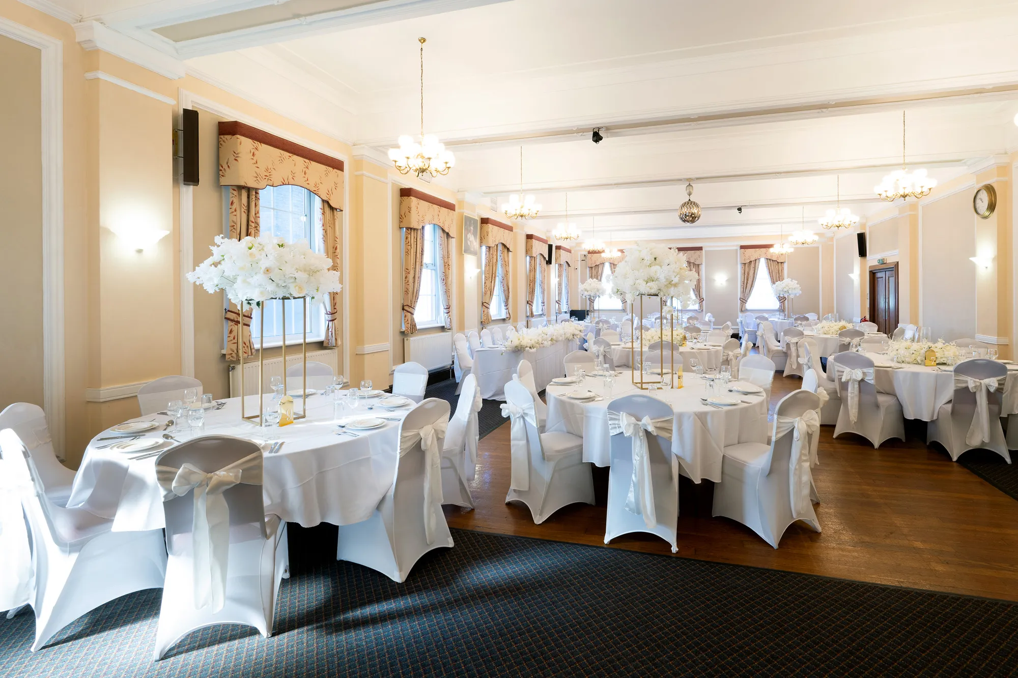 Bright wedding reception room with round tables, white chair covers, and tall white floral centerpieces.