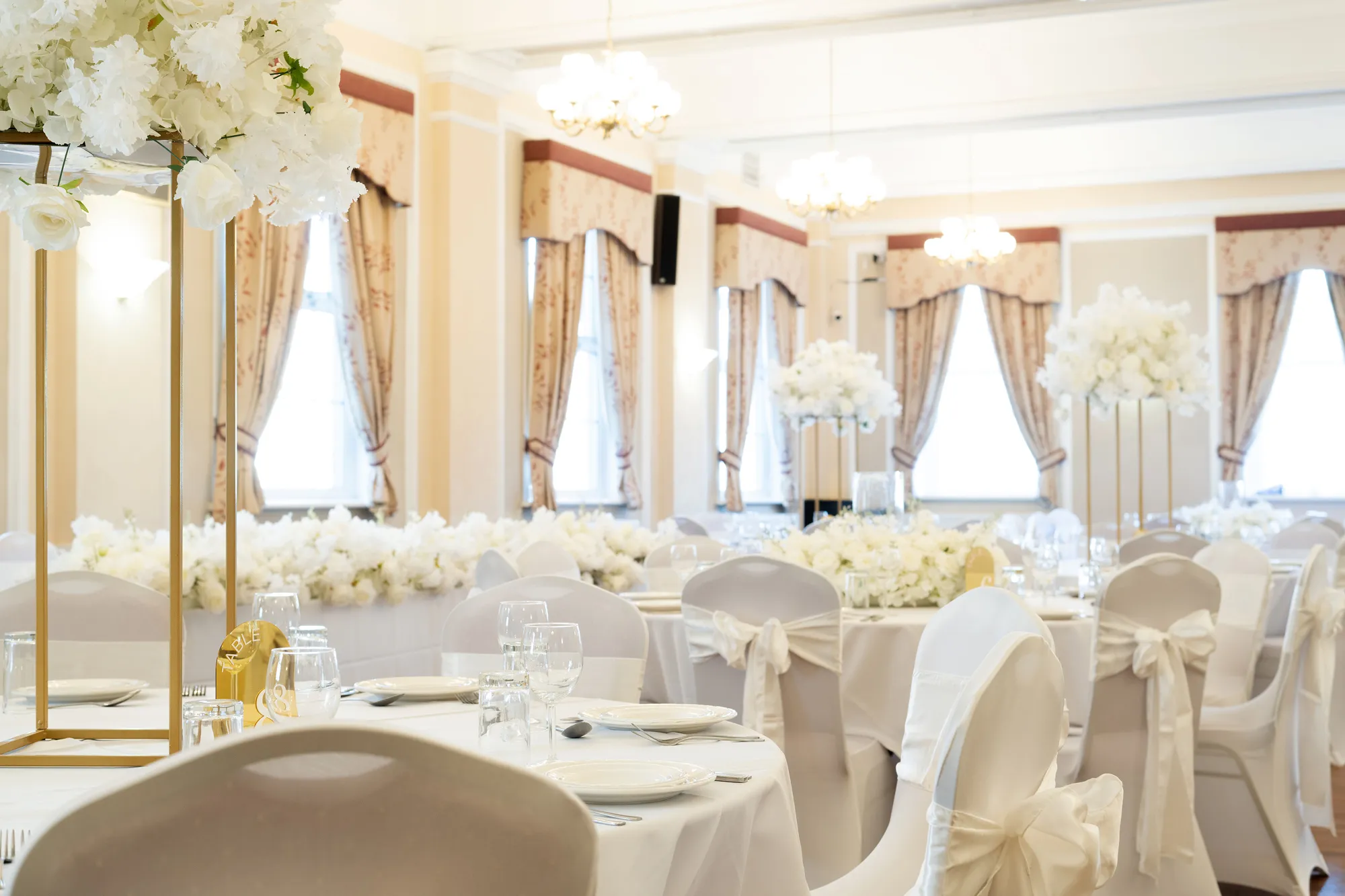 Elegant wedding reception room with round tables, white floral centerpieces, and chairs tied with white bows.