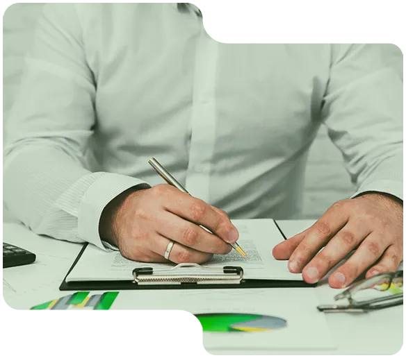A man in a white shirt writing on a clipboard with charts and glasses on the desk.