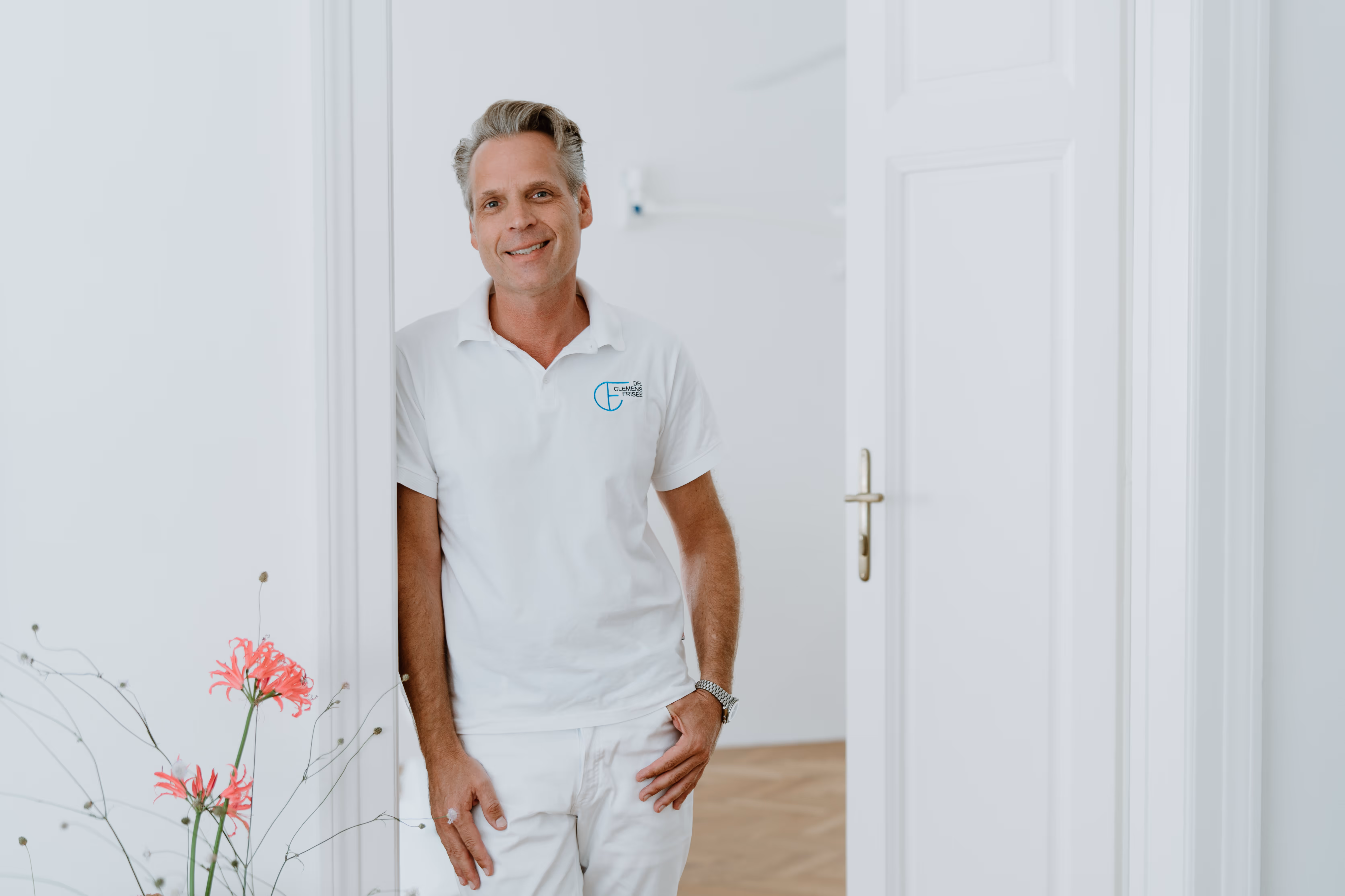 Ein lächelnder Mann in weißem Poloshirt mit Logo lehnt an einem weißen Türrahmen in einem hellen Raum.