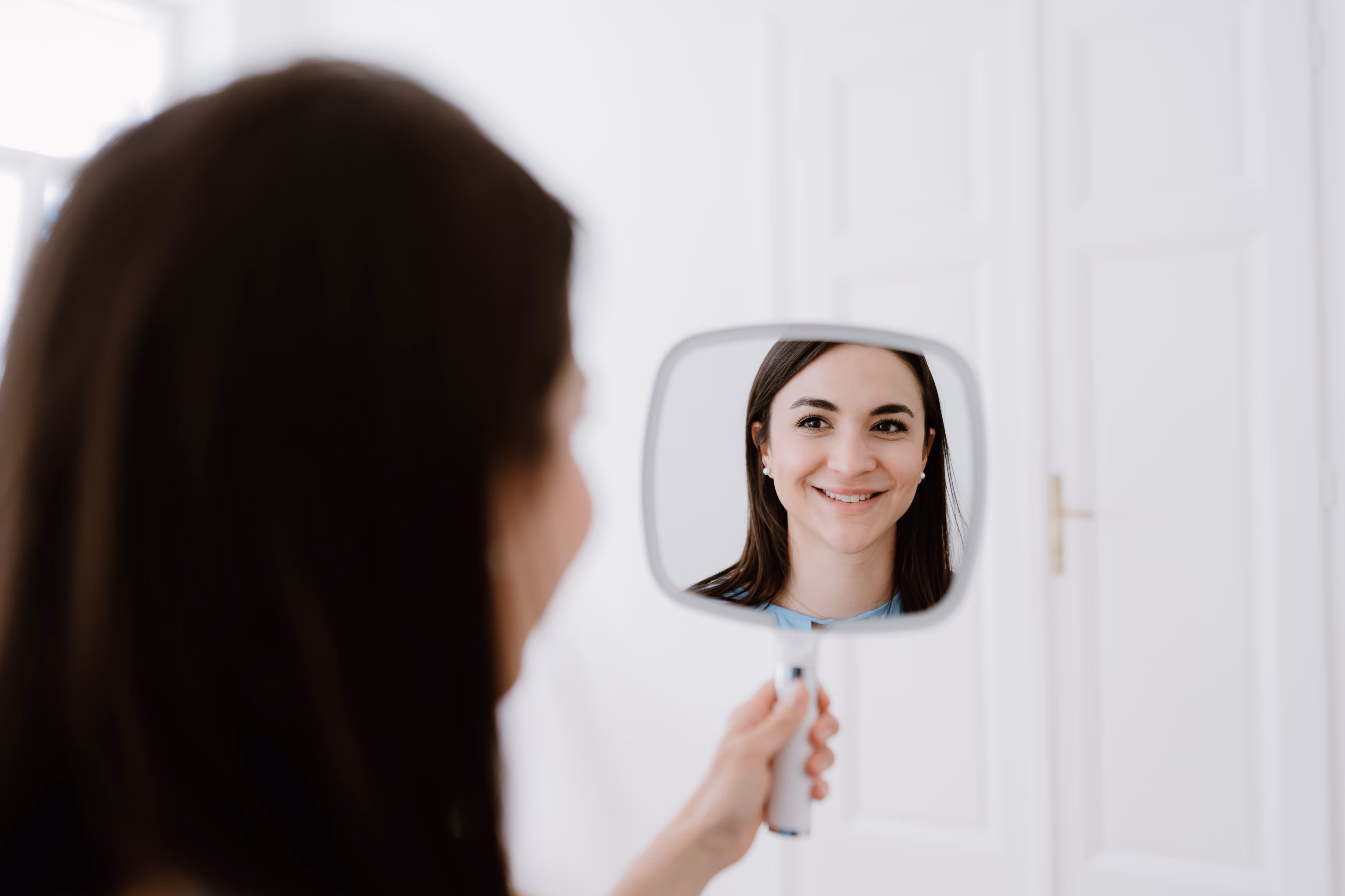 Junge Frau mit dunklen Haaren lächelt in einen Handspiegel vor weißer Tür.