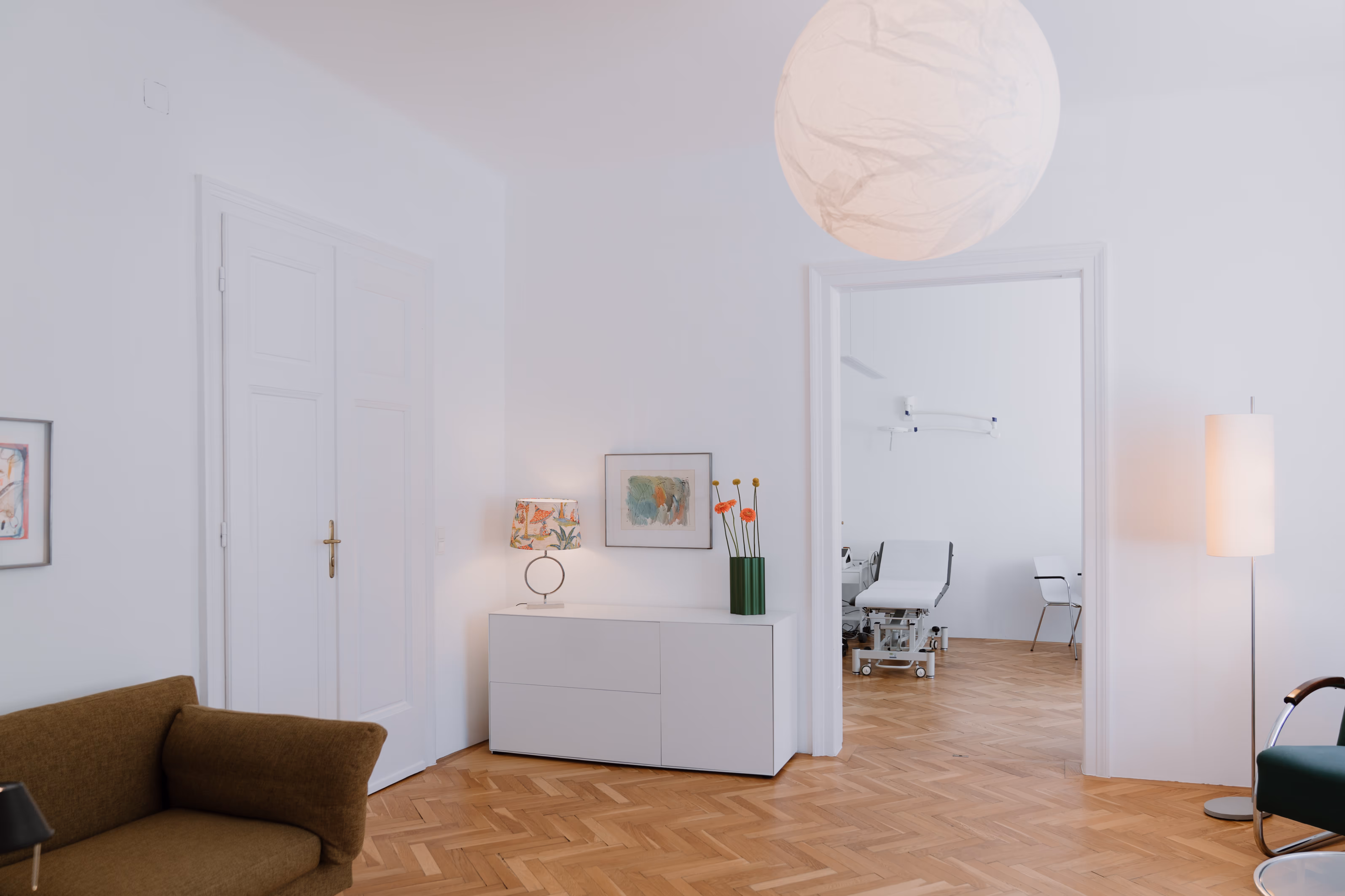 Helles Wartezimmer mit braunem Sofa, weißem Sideboard mit Lampe und Vase, sowie Blick in einen Behandlungsraum mit Untersuchungsliege.