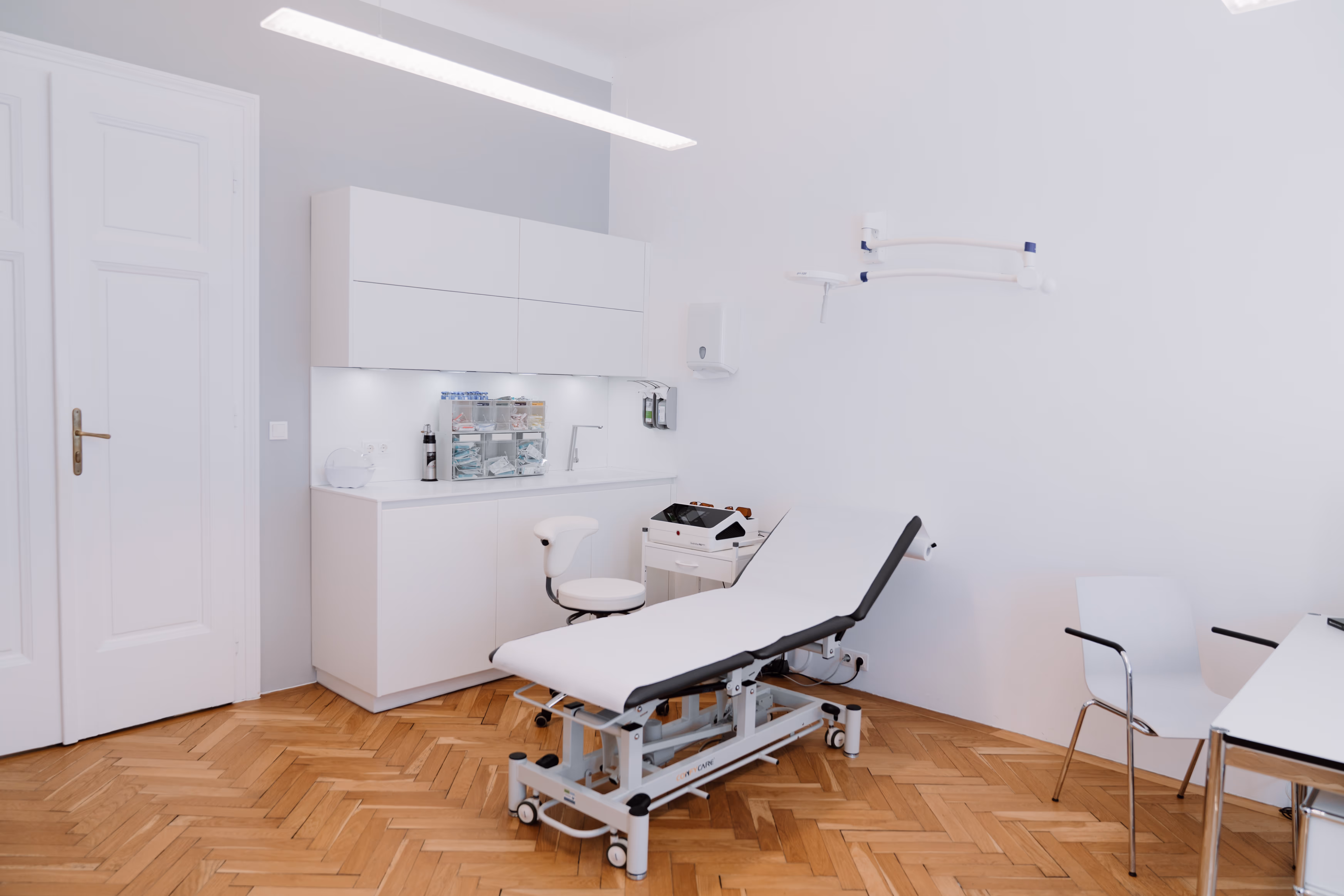 Helle, moderne Arztpraxis mit Untersuchungsliege, Stuhl, weißen Schränken und Holzboden.