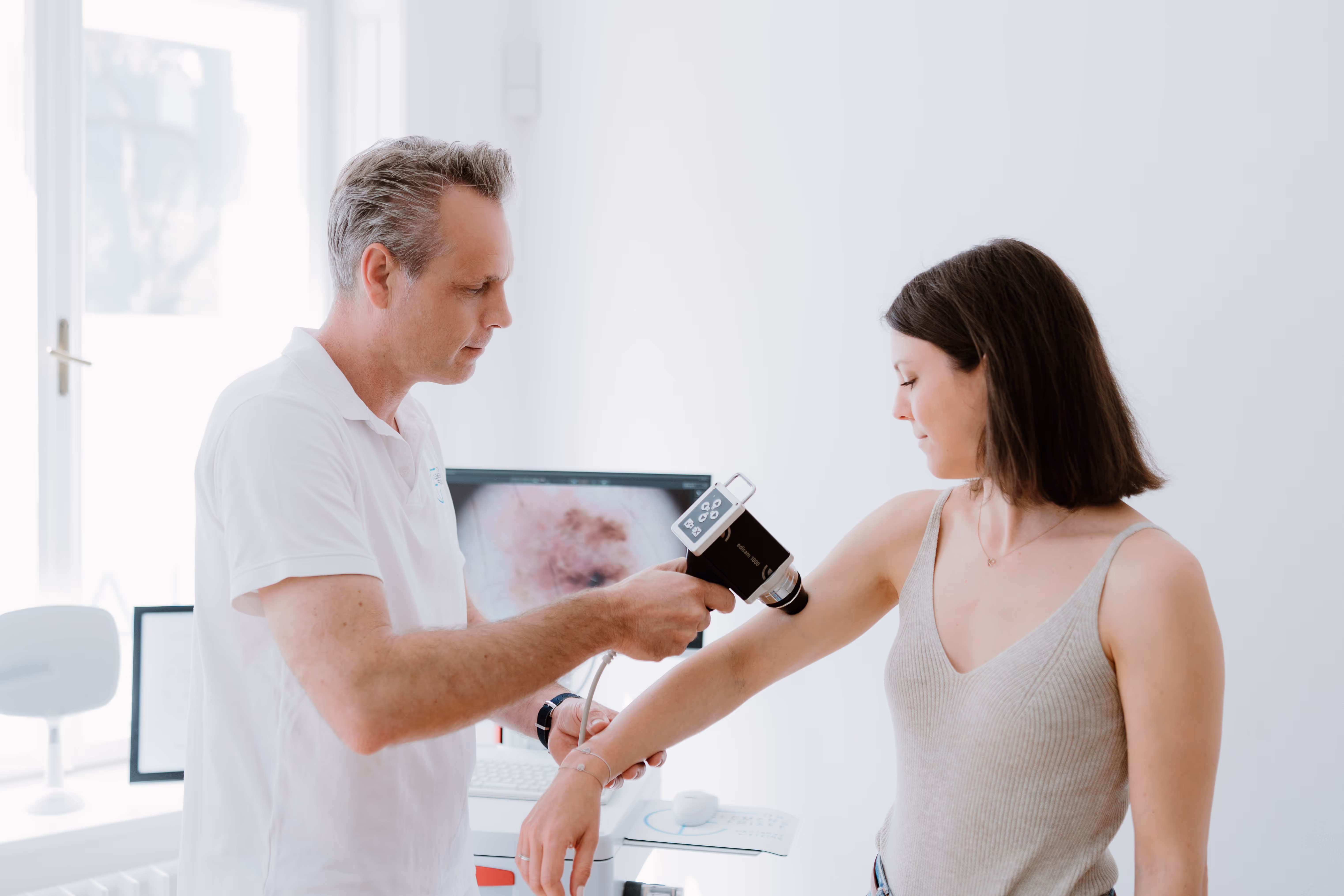 Dr. Clemens Frisee untersucht den Arm einer Frau mit einem Dermatoskop in einer hellen Praxis.