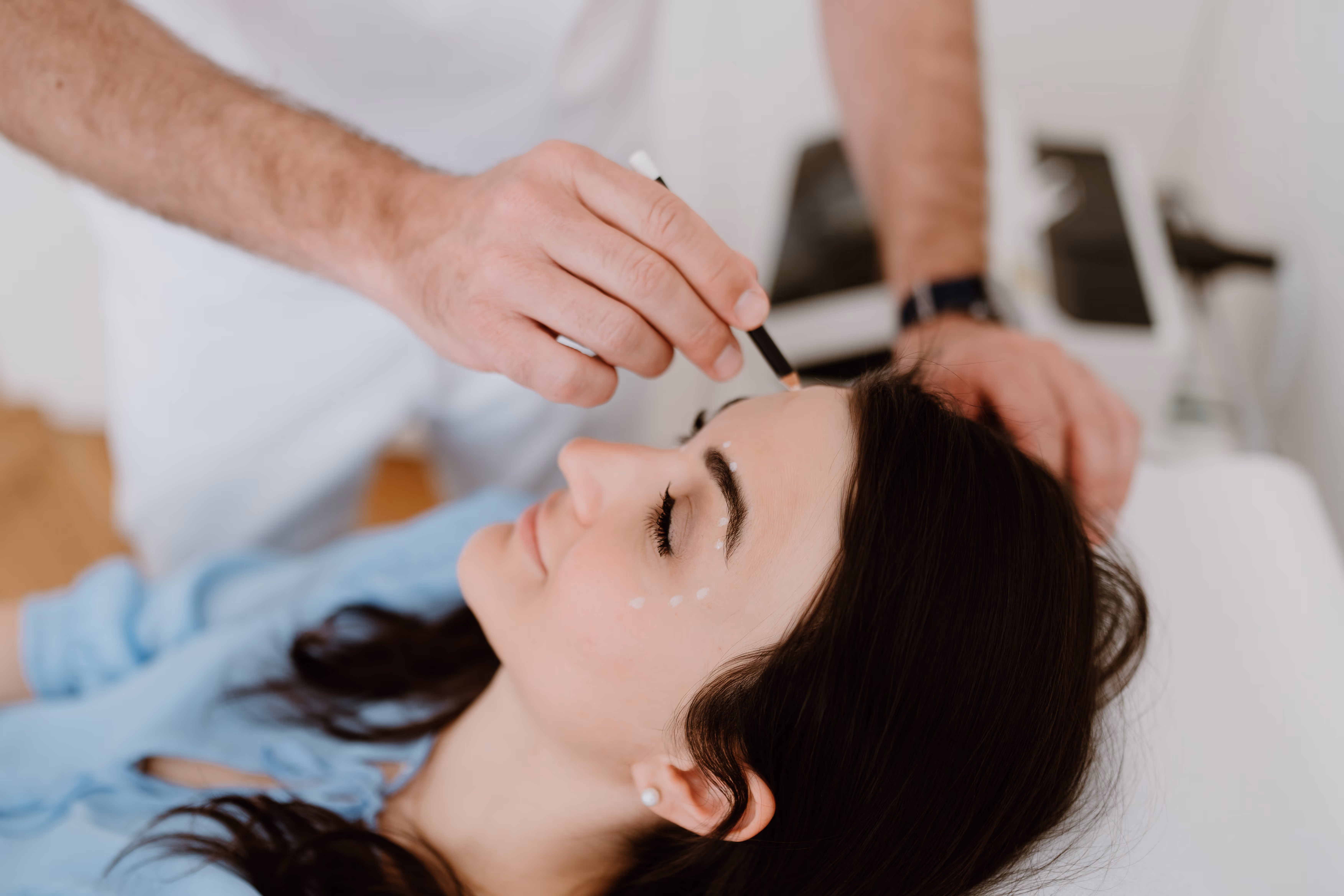 Frau liegt entspannt mit geschlossenen Augen, während Dr. Clemens Frisee weiße Markierungen auf ihrer Stirn setzt.