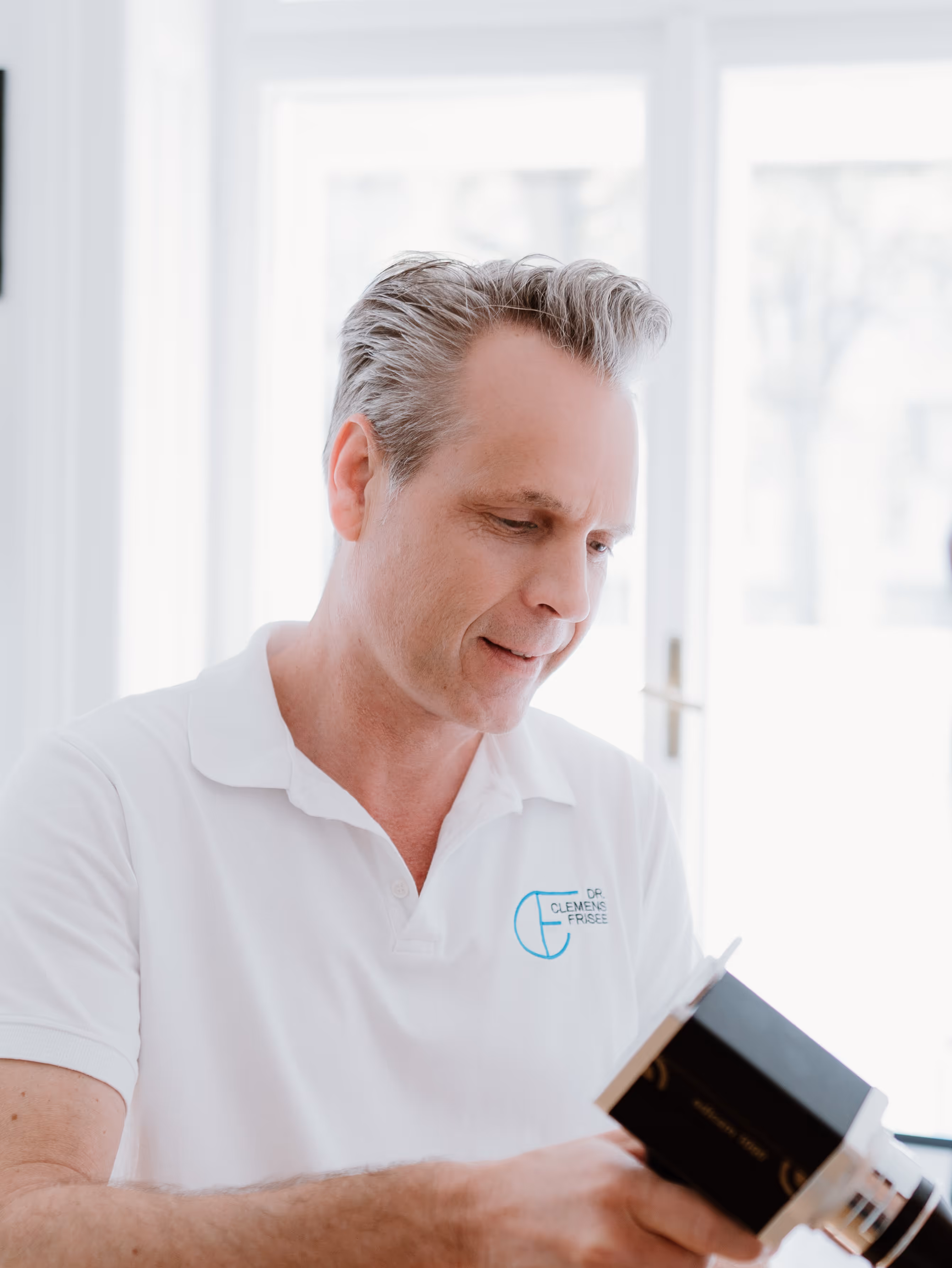 Mann im weißen Polohemd mit Aufschrift Dr. Clemens Frisee liest ein schwarzes Buch in einem hellen Raum.