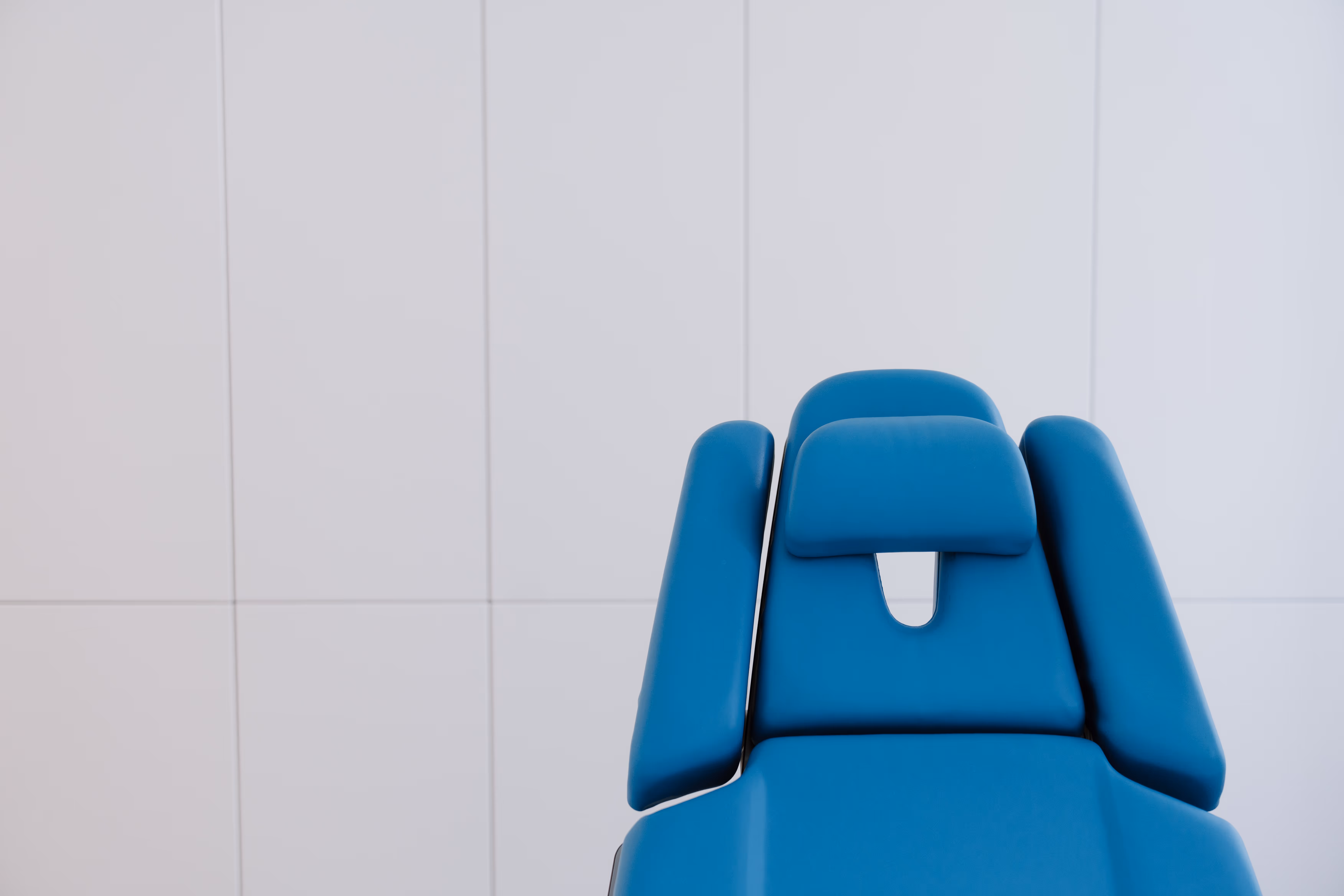 Blauer Behandlungsstuhl vor neutraler, weißer Wand mit Kacheln.