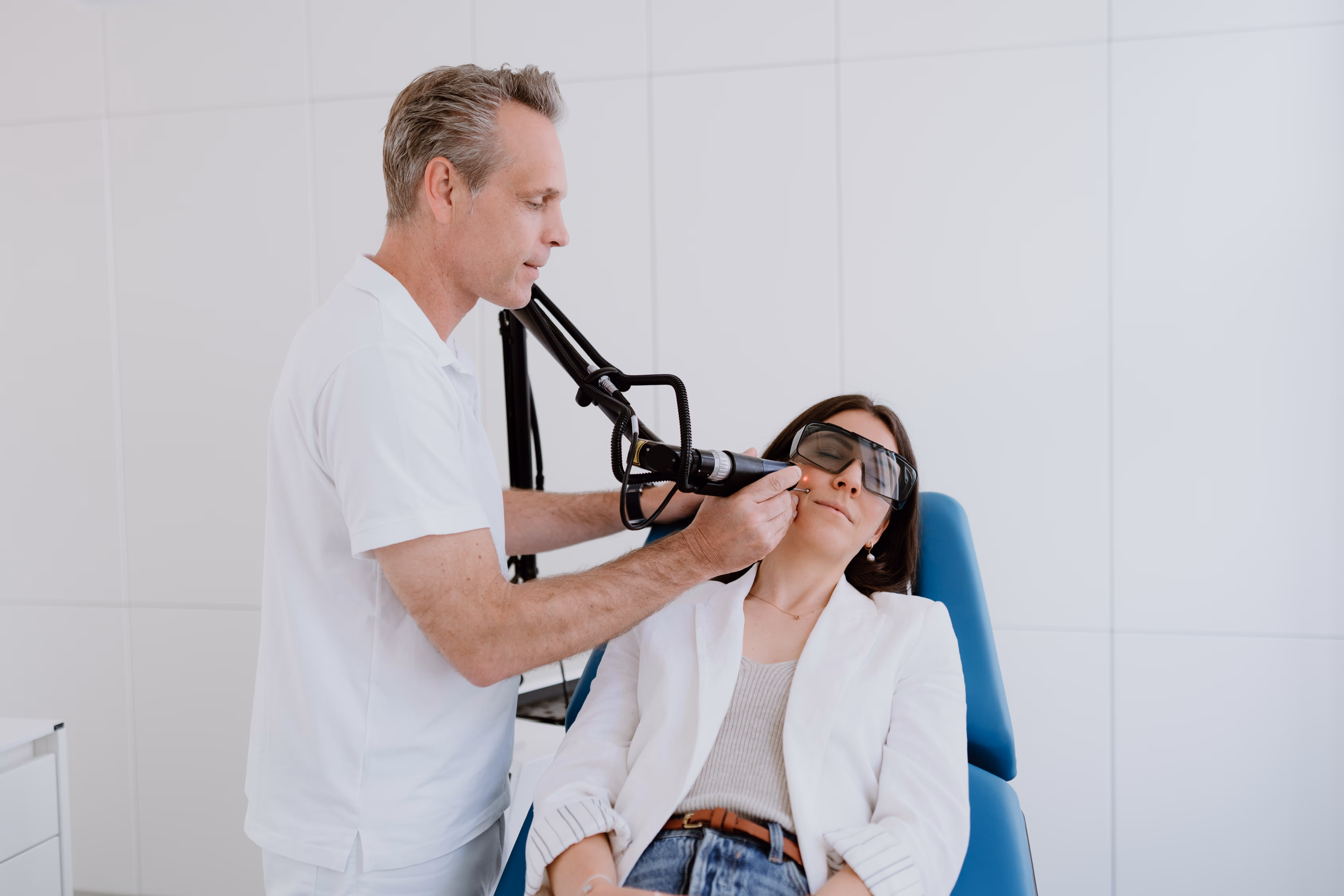 Dr. Clemens Frisee führt eine Laser-Hautbehandlung an der Wange einer Frau durch, die eine Schutzbrille trägt, während sie auf einem Behandlungsstuhl sitzt.
