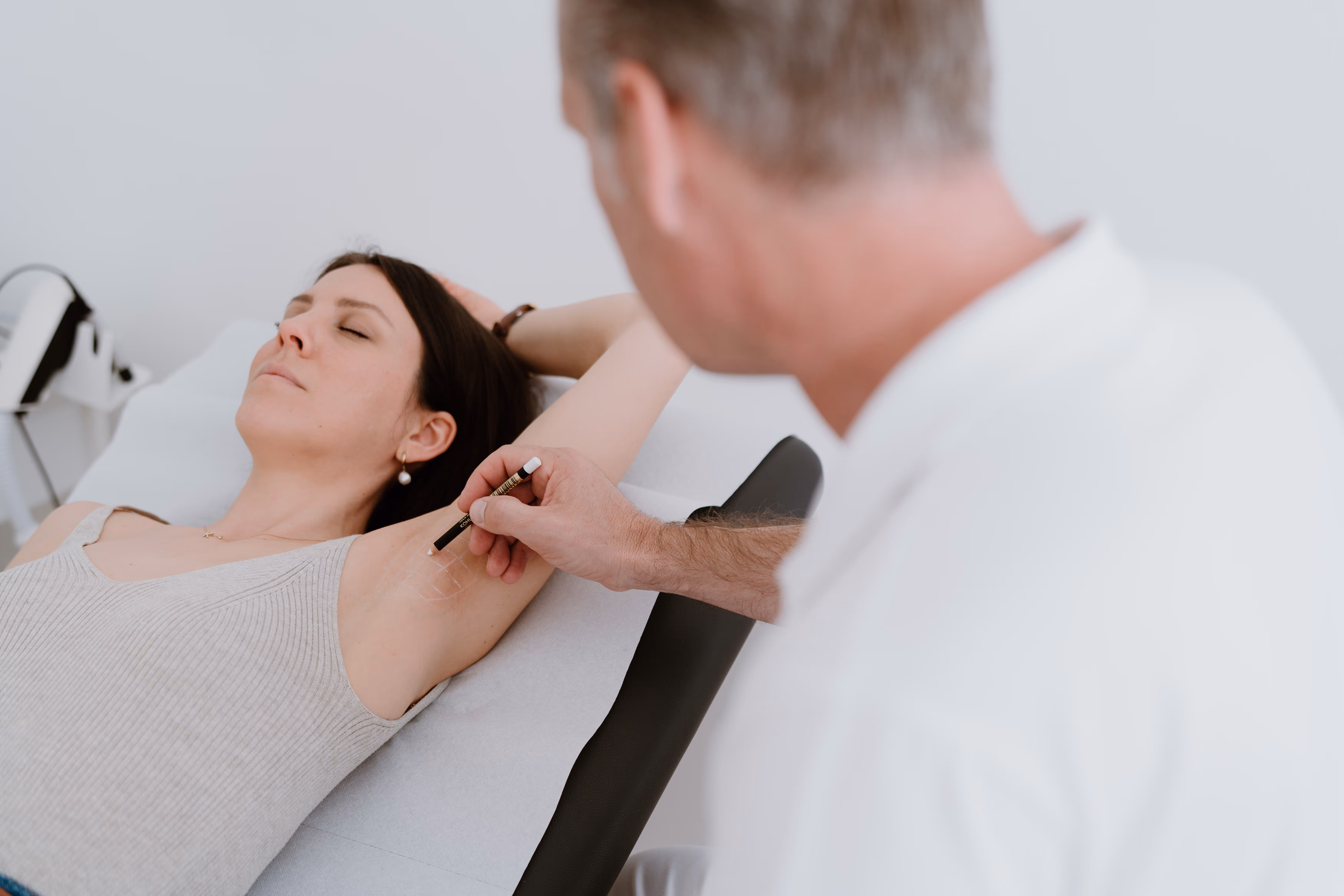Arzt markiert mit einem Stift die Achselhöhle einer entspannten liegenden Frau für eine medizinische Behandlung.