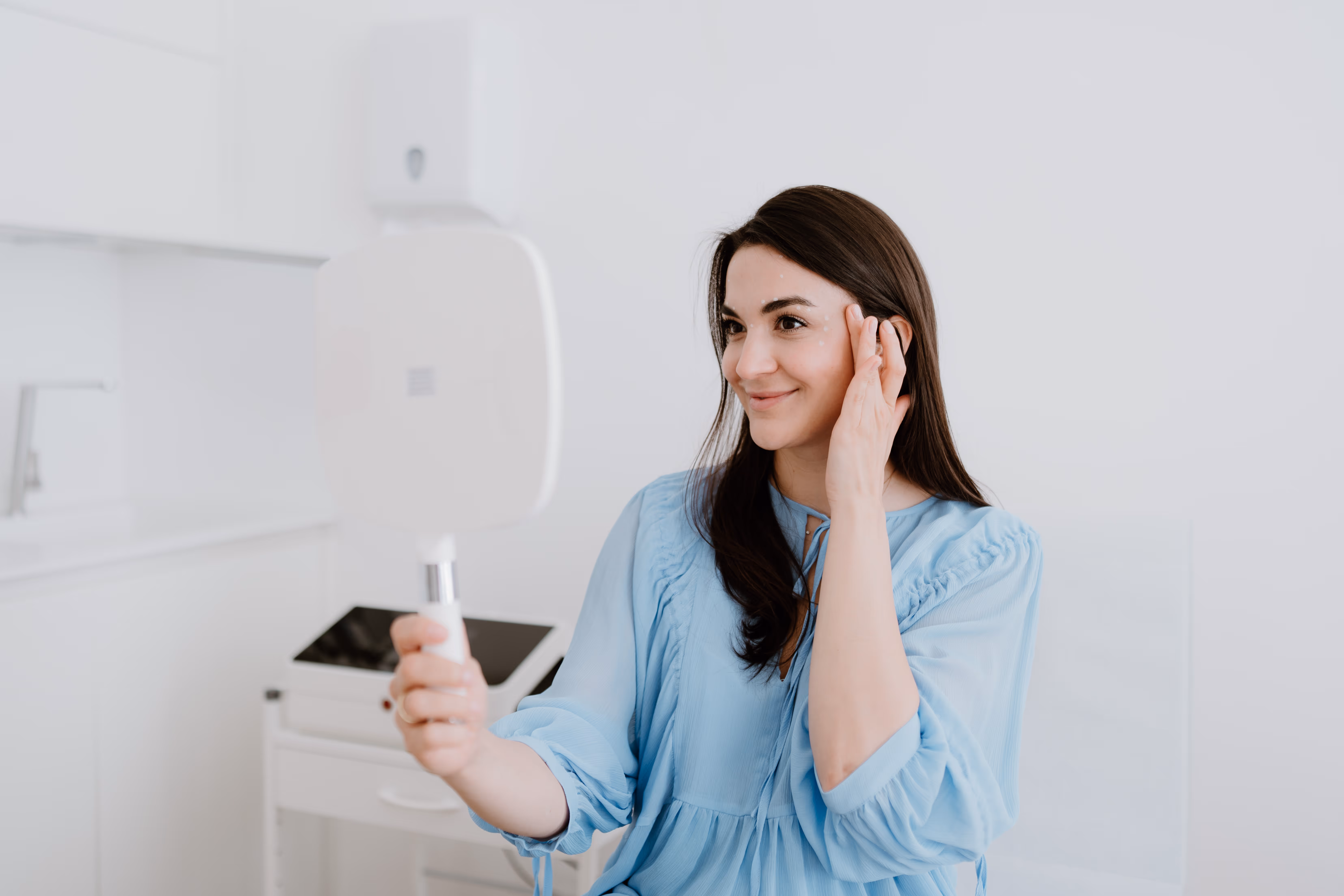 Frau mit dunkelbraunen Haaren in blauem Oberteil betrachtet ihr Gesicht in einem Handspiegel in einem hellen Raum.