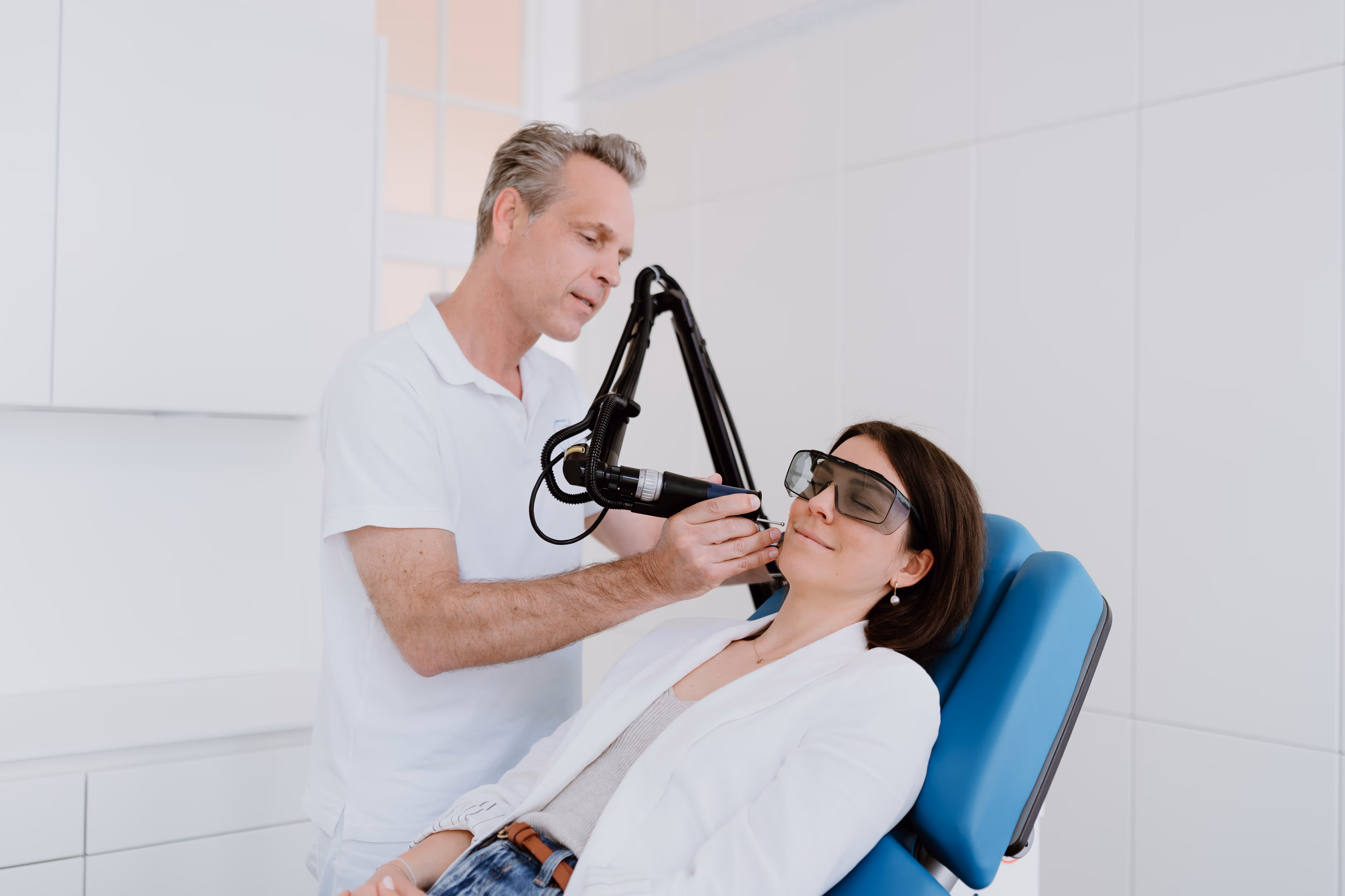 Hautarzt Dr. Clemens Frisee führt eine Hautbehandlung mit einem Lasergerät an einem entspannten Patienten mit Schutzbrille in einer hellen Klinik durch.