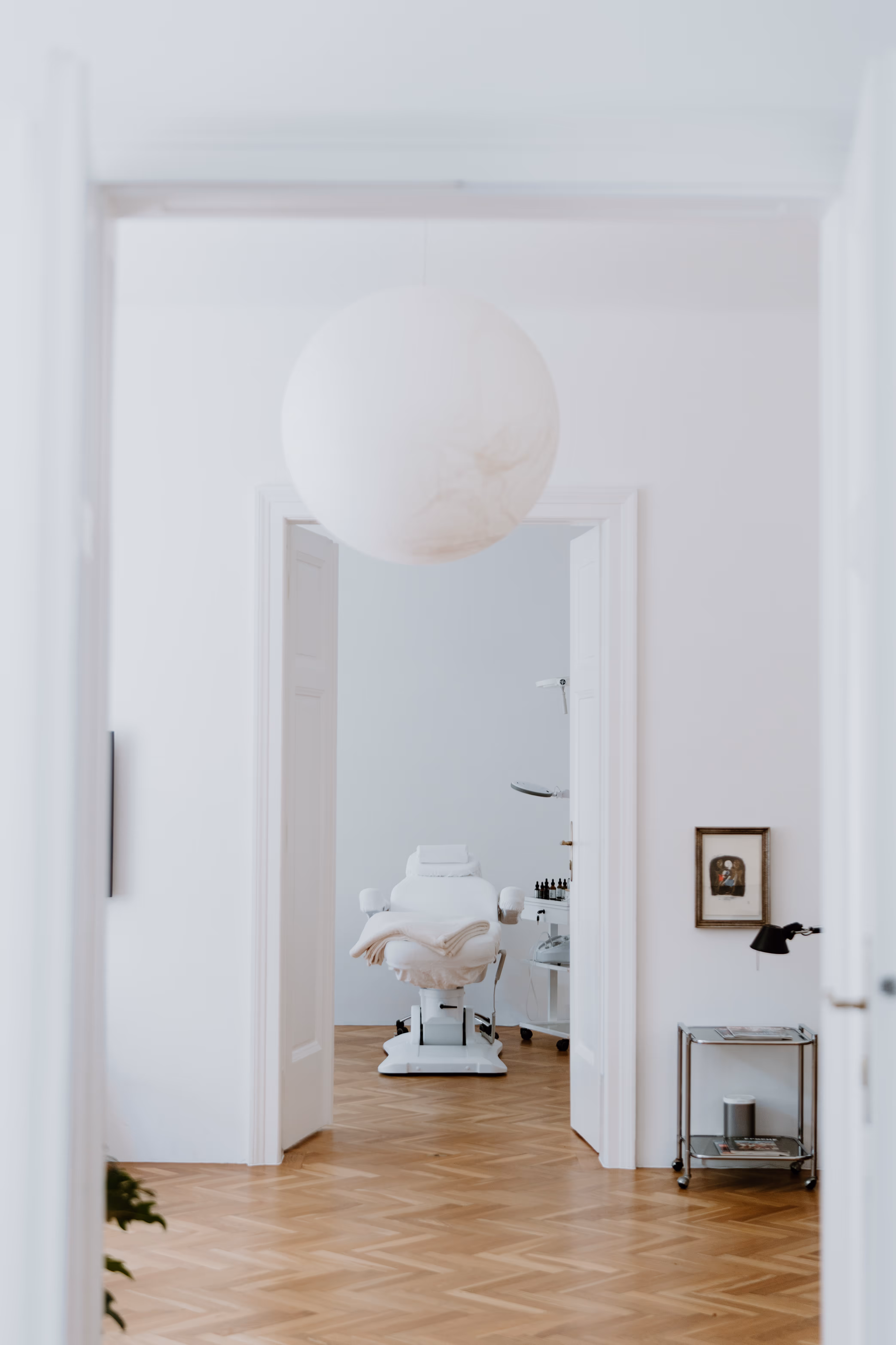 Behandlungszimmer mit weißem, gepolstertem Behandlungsstuhl, hölzernem Parkettboden und einer runden weißen Deckenleuchte.