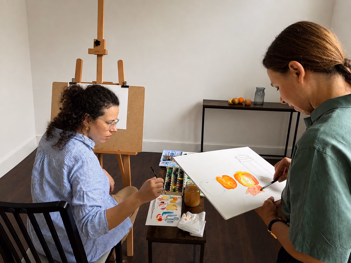 Two women painting fruit still life with watercolors in a simple room with wooden floor.