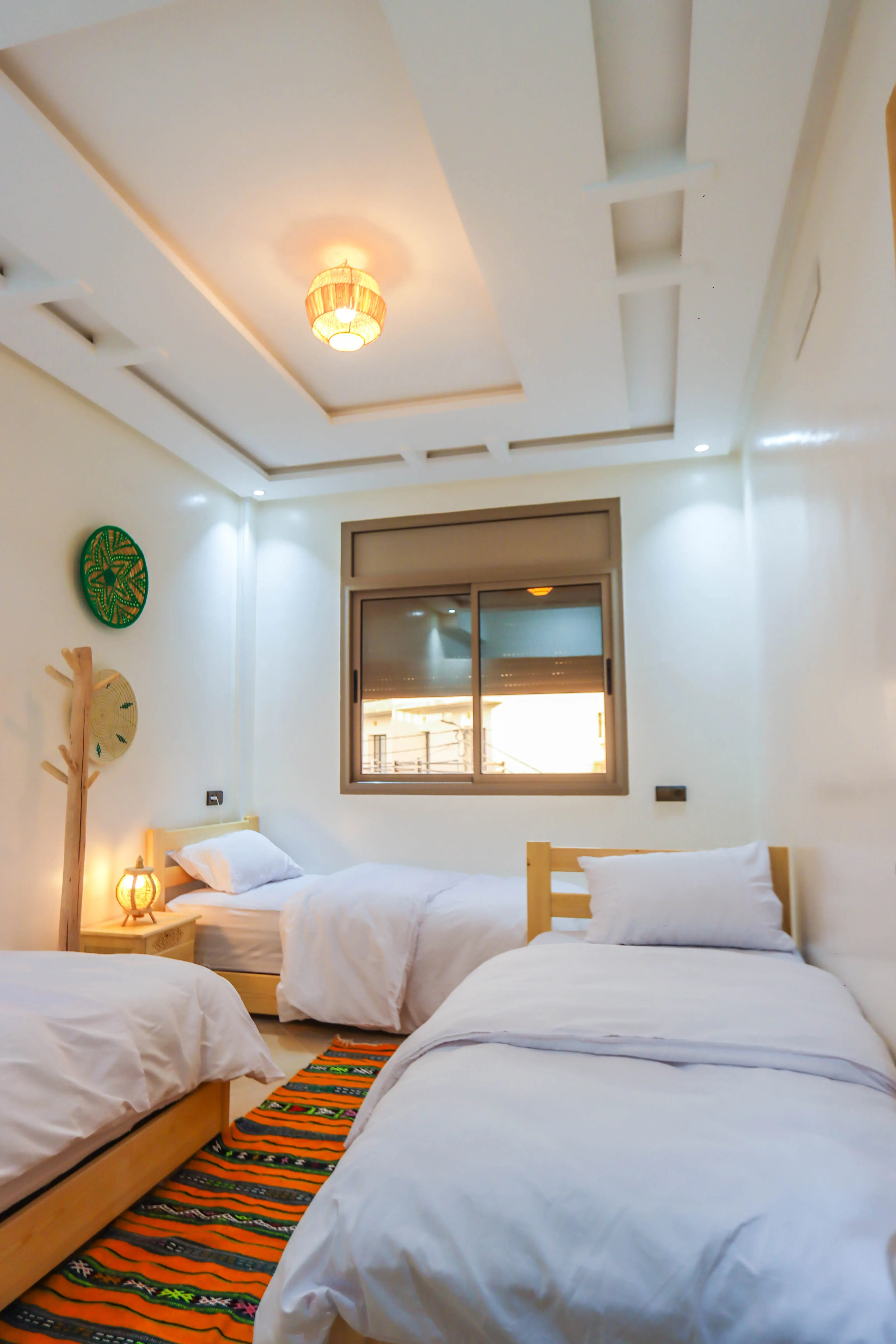 Bright bedroom with three single beds, white bedding, a window, and a colorful rug on the floor.