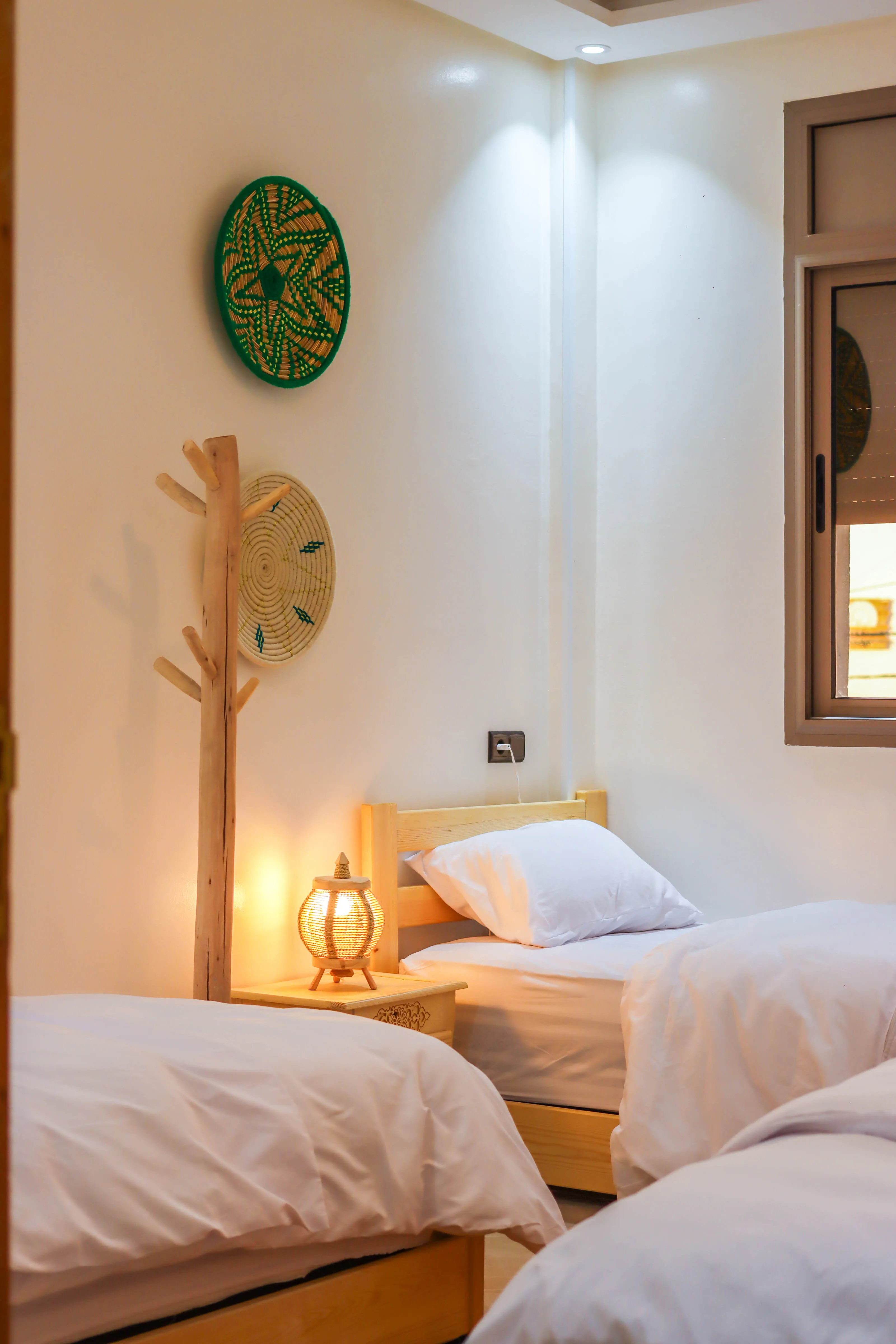 Cozy bedroom with two single wooden beds, white bedding, a nightstand with a lit lamp, and decorative wall hangings.