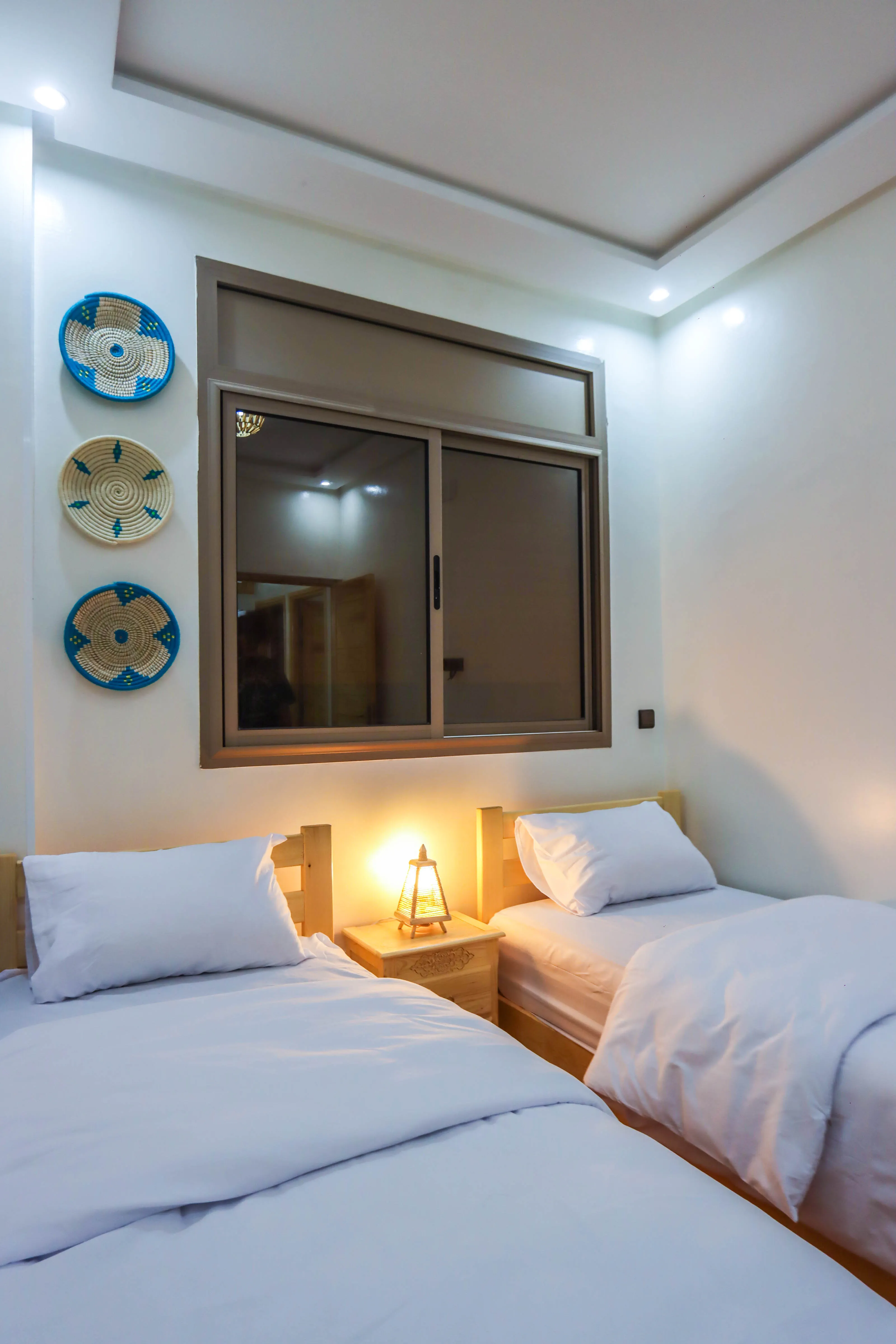 Simple bedroom with two single beds, white bedding, a wooden nightstand, and lit lamp under a window.