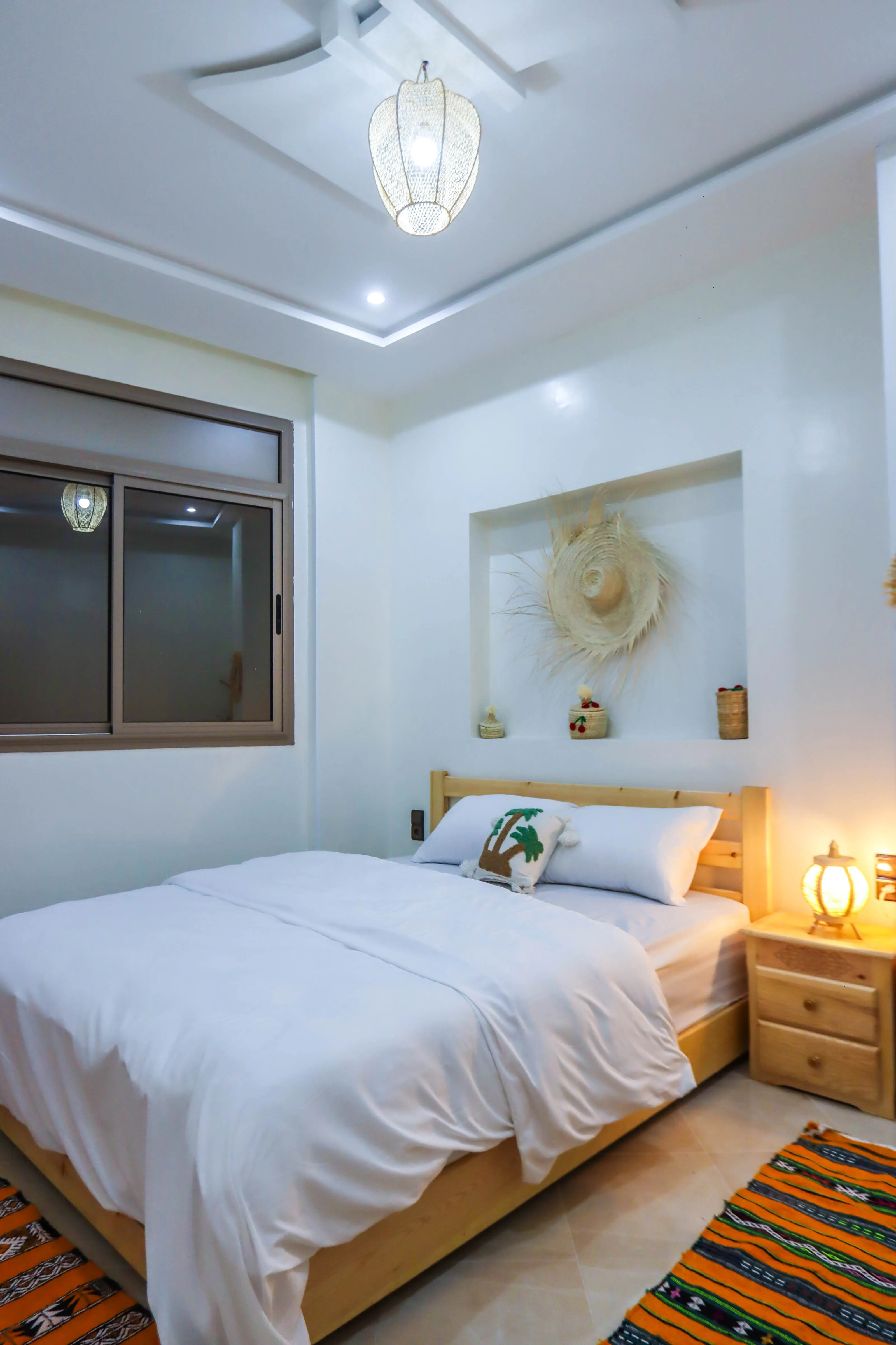 Cozy bedroom with a wooden bed, white bedding, a palm tree pillow, and warm lighting.