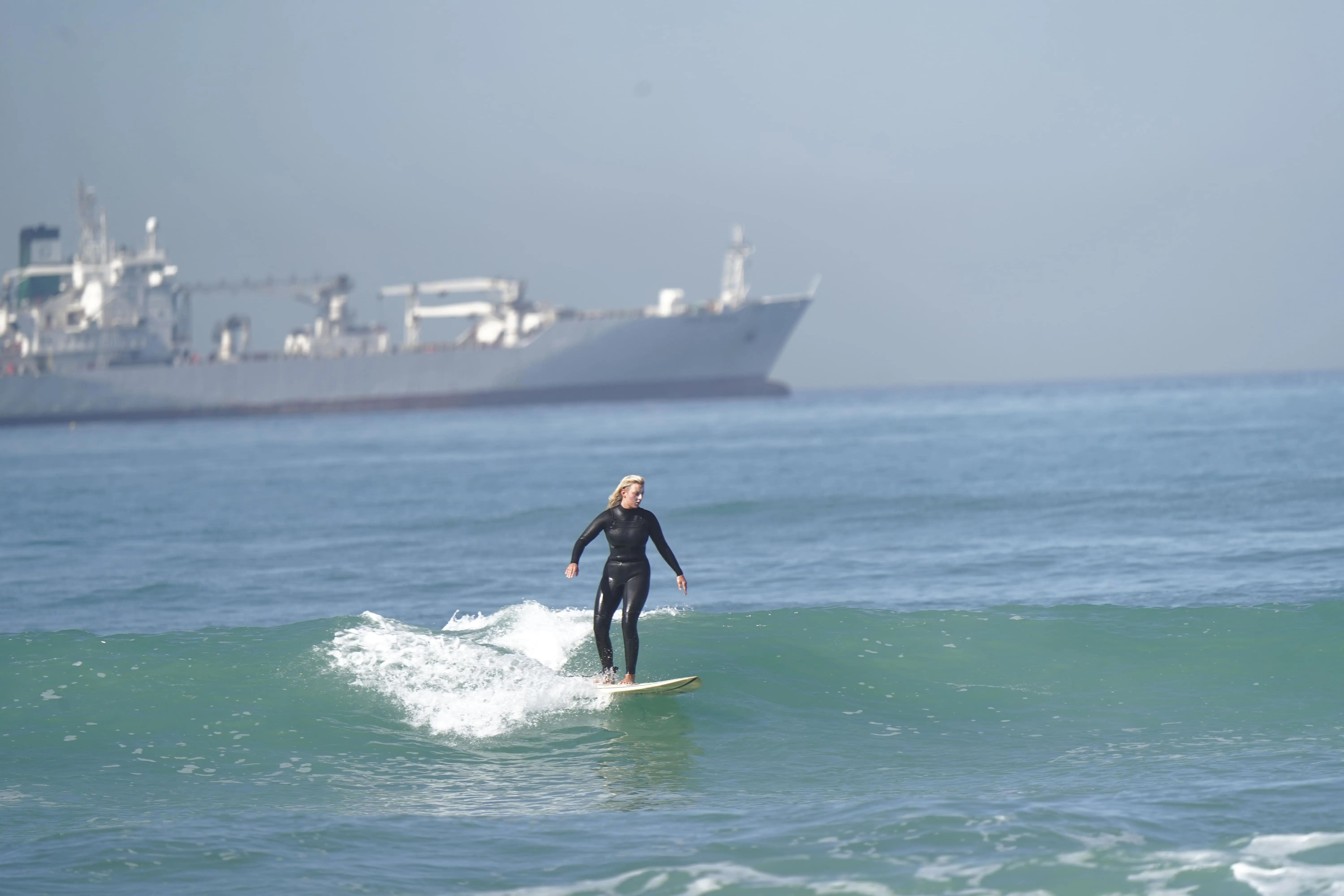 Person in a black wetsuit surfing a small wave with a large ship in the background.