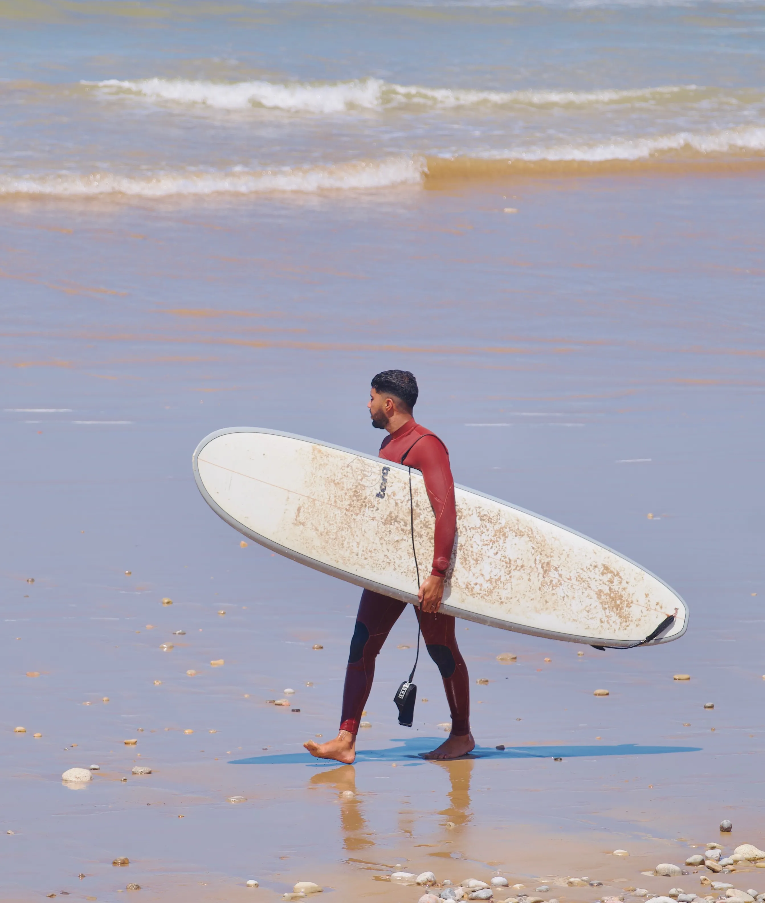 Man in a red wetsuit carrying a surfboard and walking barefoot on a sandy beach with small rocks.