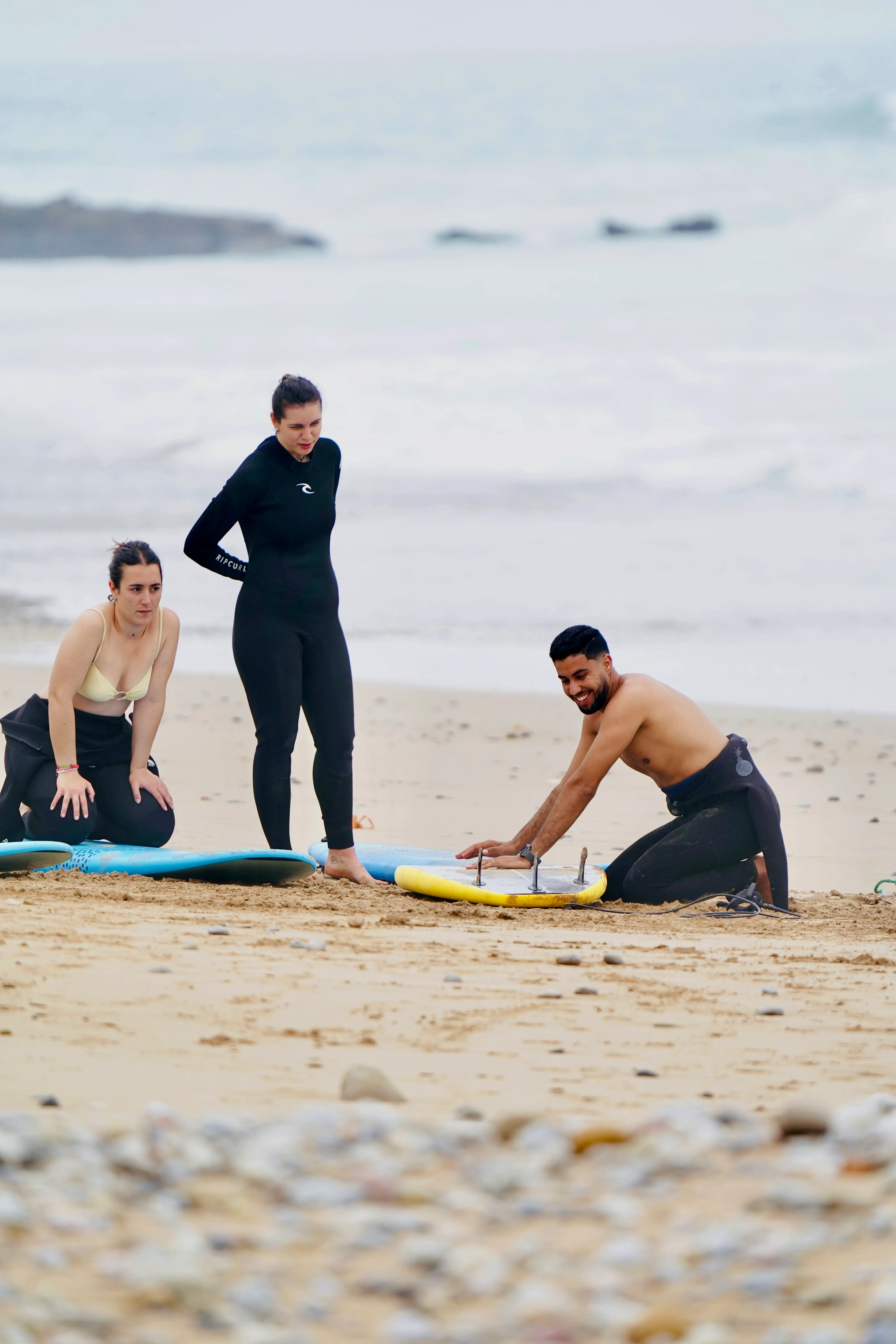 Three people on a sandy beach with surfboards, one man kneeling adjusting a surfboard fin.