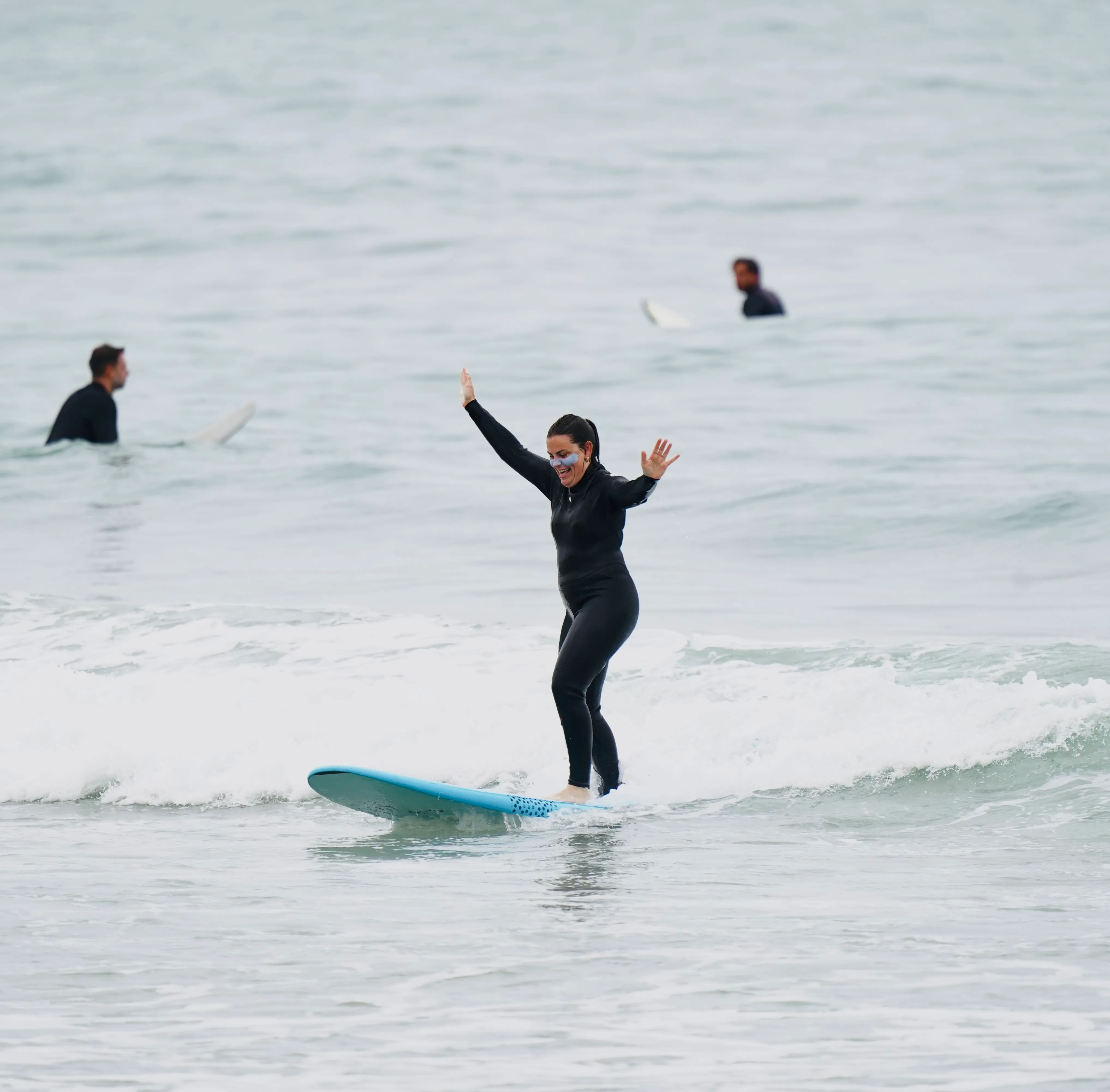 Woman balancing on a blue surfboard riding a small wave, wearing a black wetsuit and face paint.