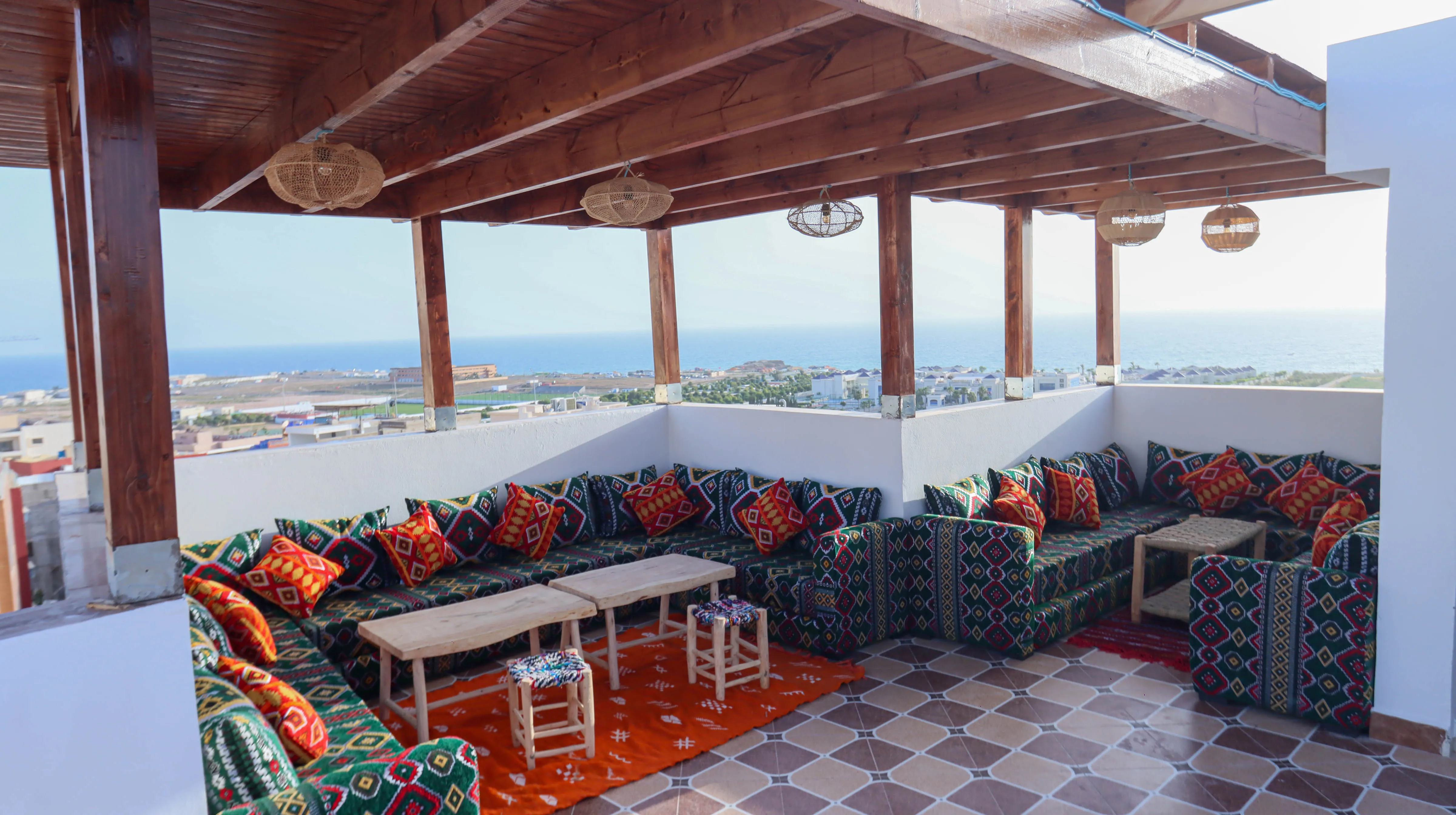 Covered rooftop lounge with patterned cushions, wooden tables, stools, and ocean view in the background.