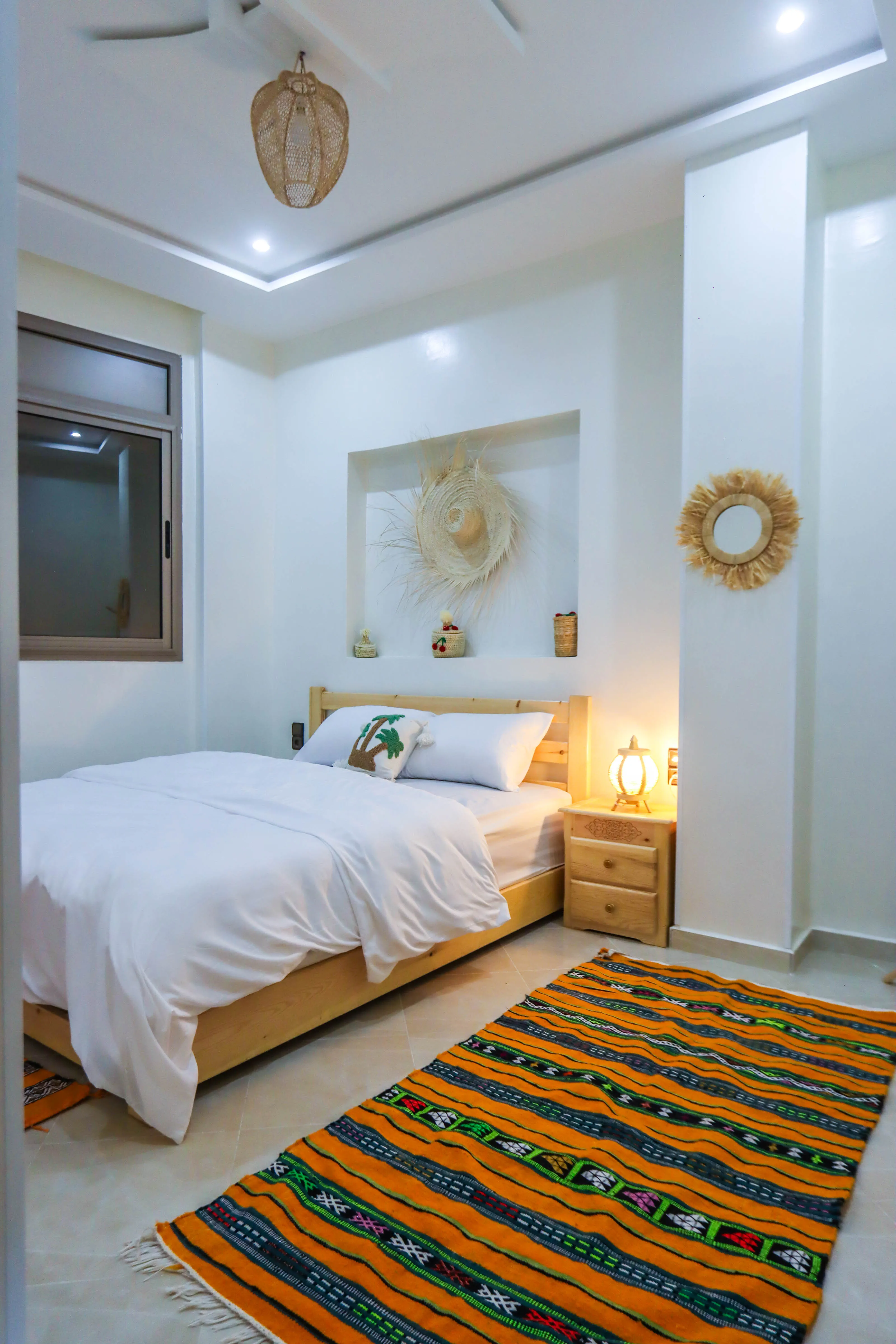 Bright bedroom with wooden bed, white bedding, colorful striped rug, and woven wall decorations.