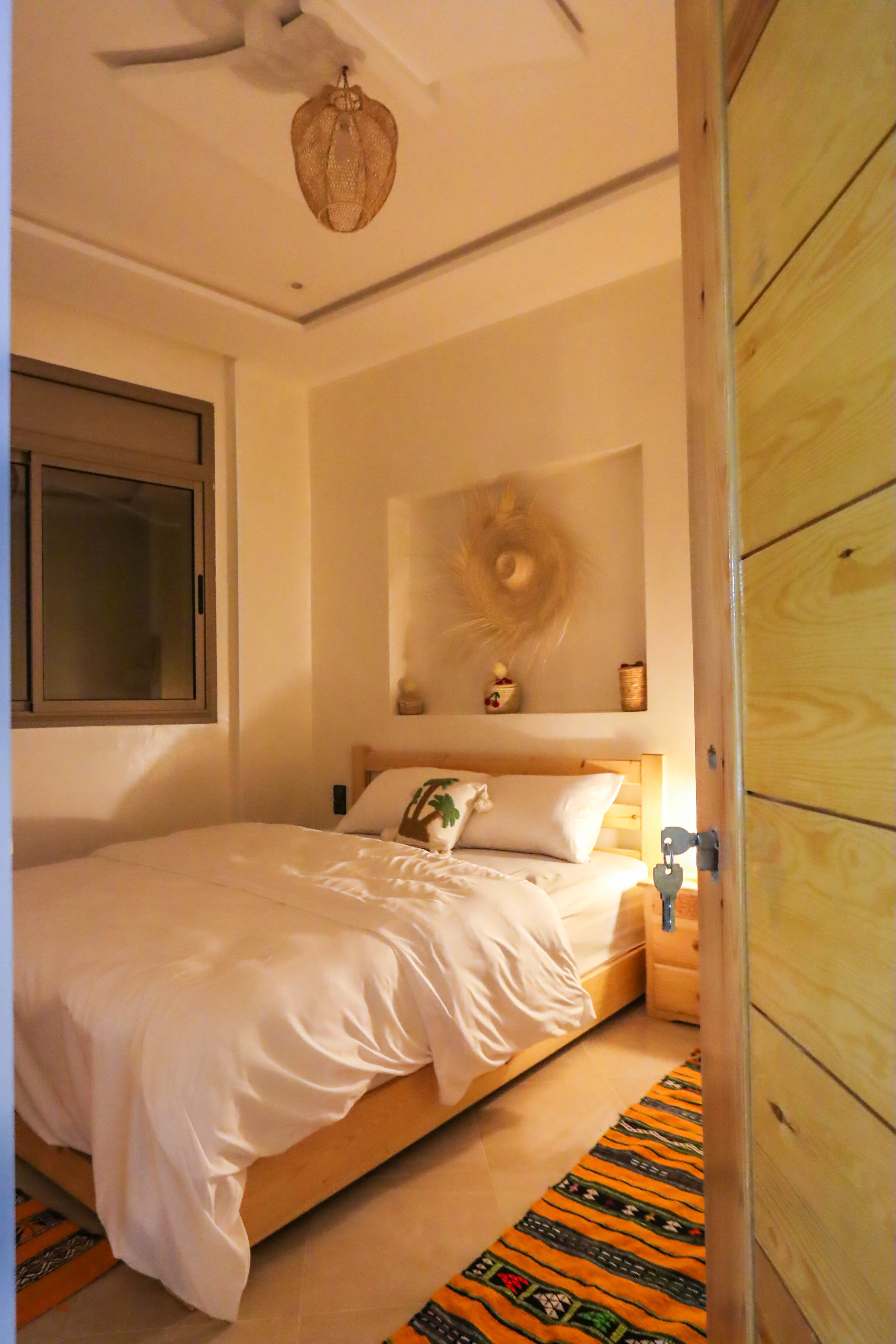 Cozy bedroom with wooden bed, white bedding, a palm tree pillow, and woven wall decoration.