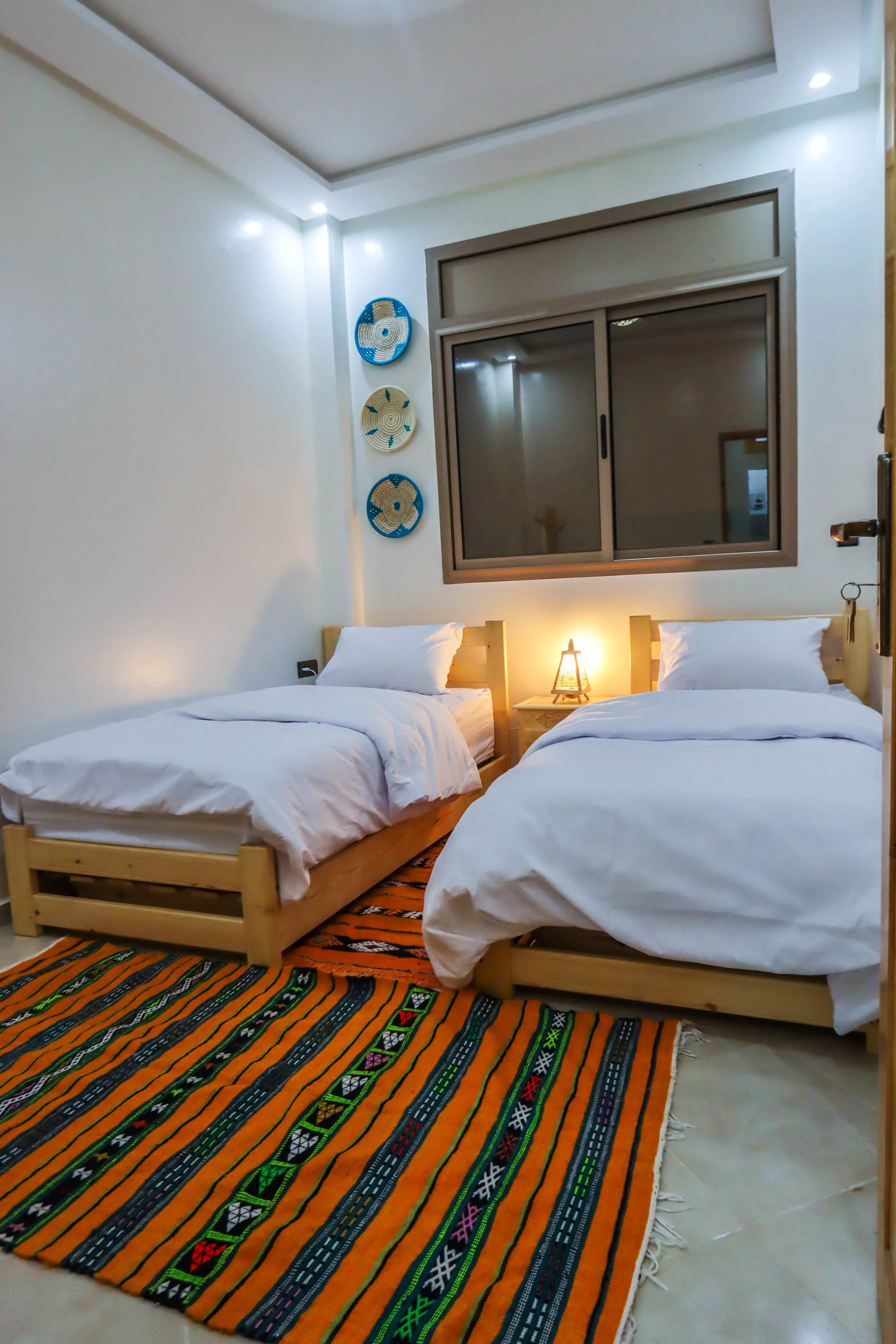 Minimalist bedroom with two twin beds, white bedding, a nightstand with a lamp, and a colorful patterned rug.