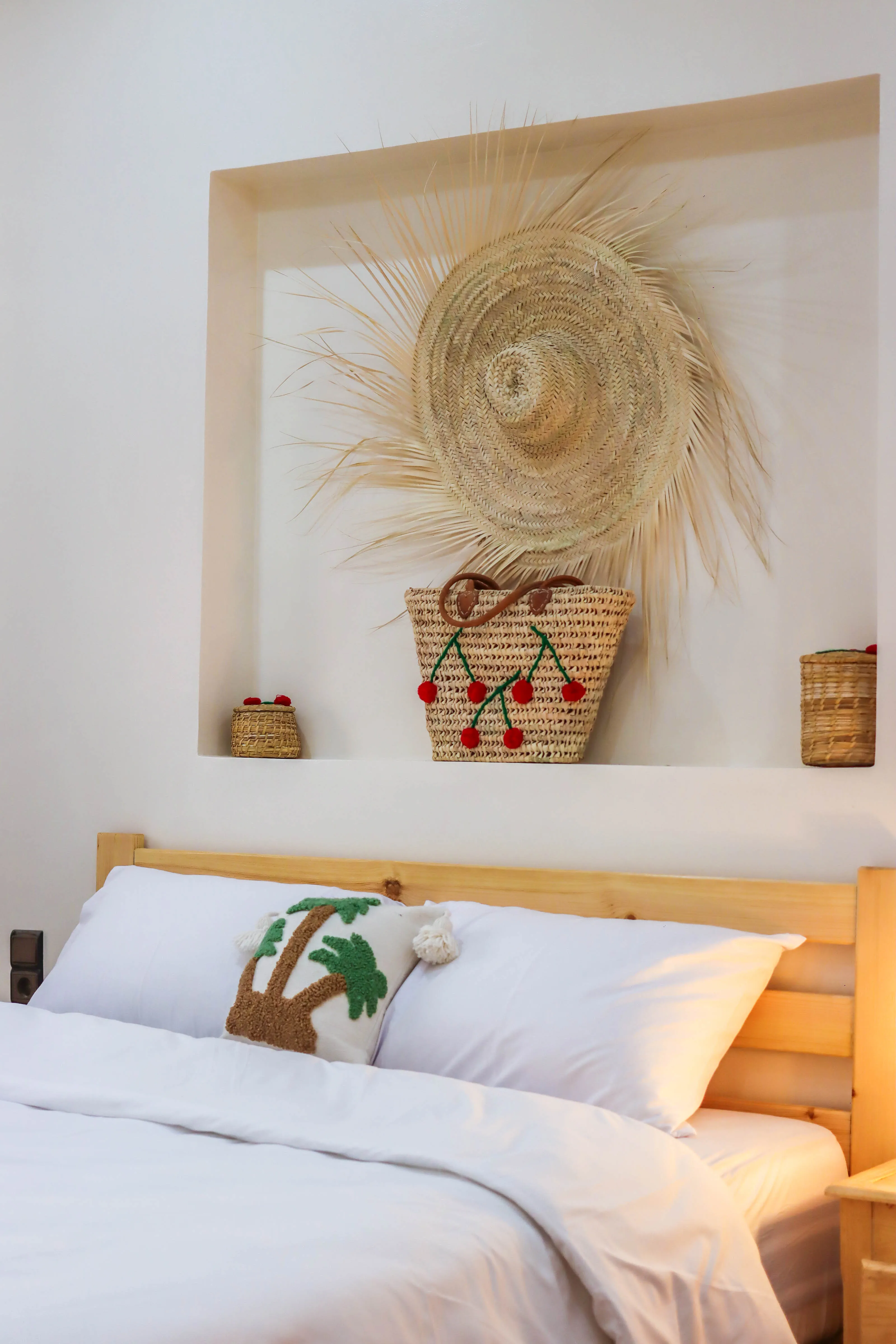Bed with white linens and palm tree pillow in front of wall niche with woven straw hat and baskets.