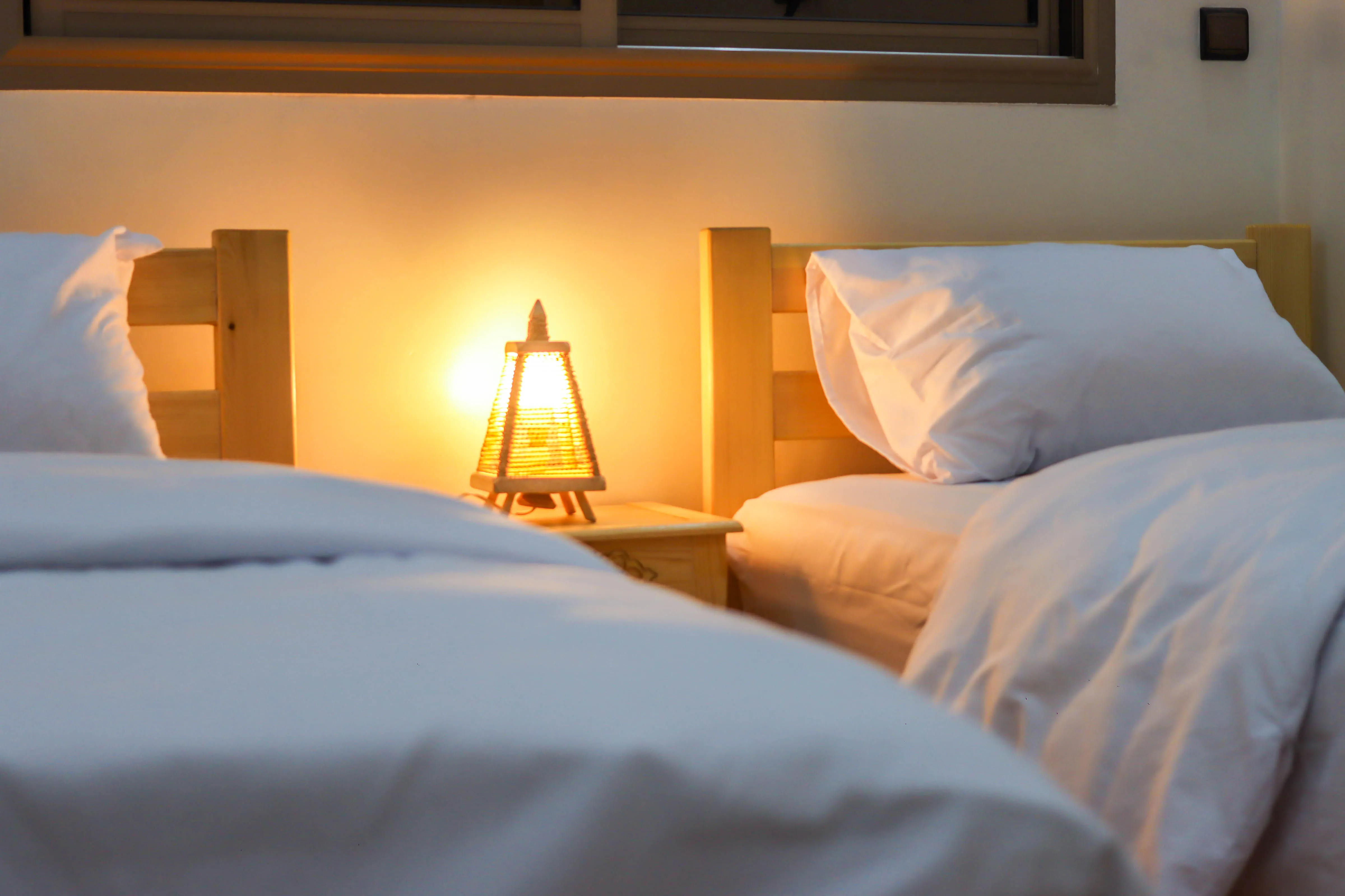 Two wooden twin beds with white bedding and a lit lamp on a nightstand between them.