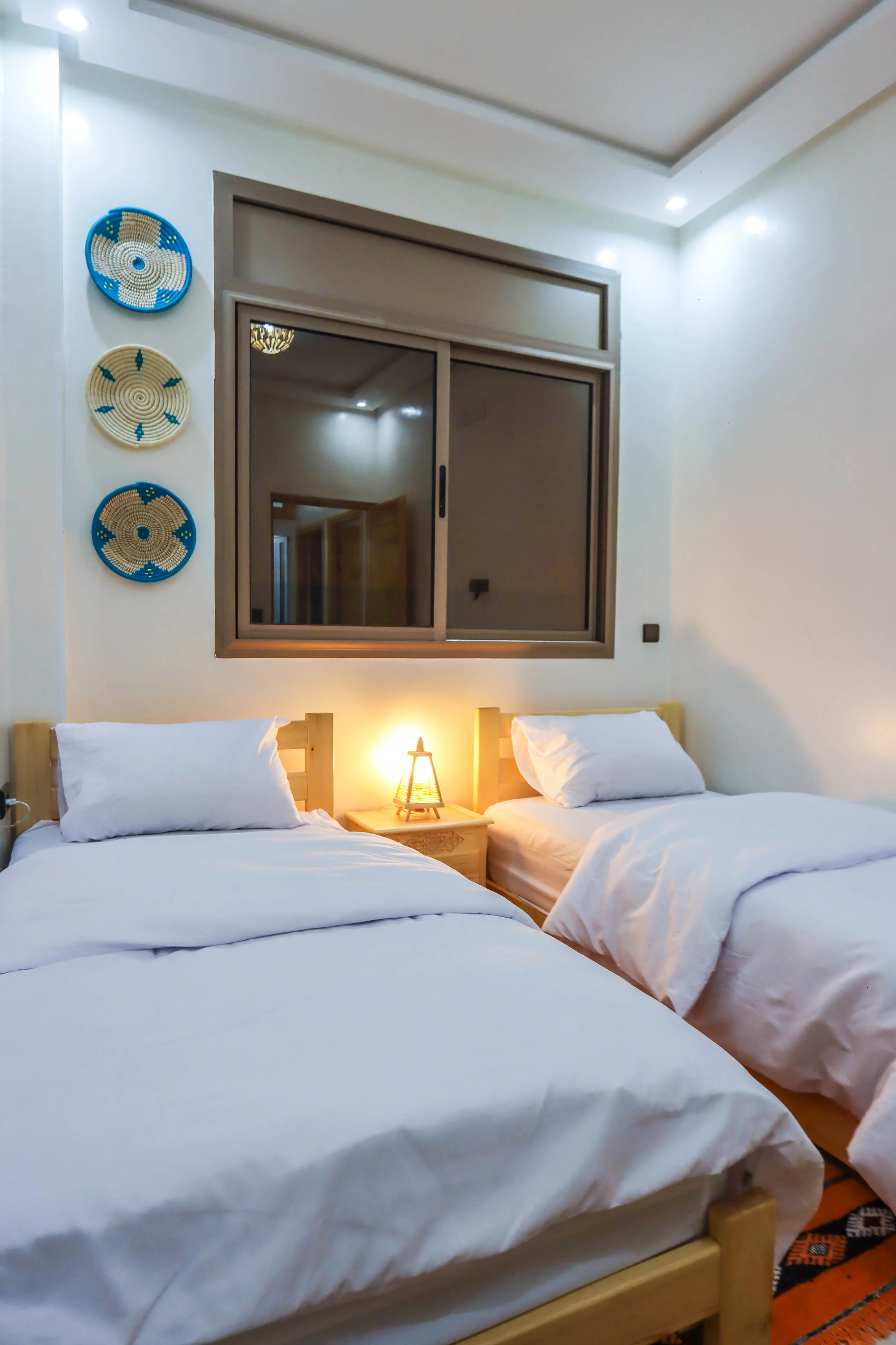 Cozy bedroom with two single beds, white bedding, a nightstand with a lamp and decorative woven wall plates.