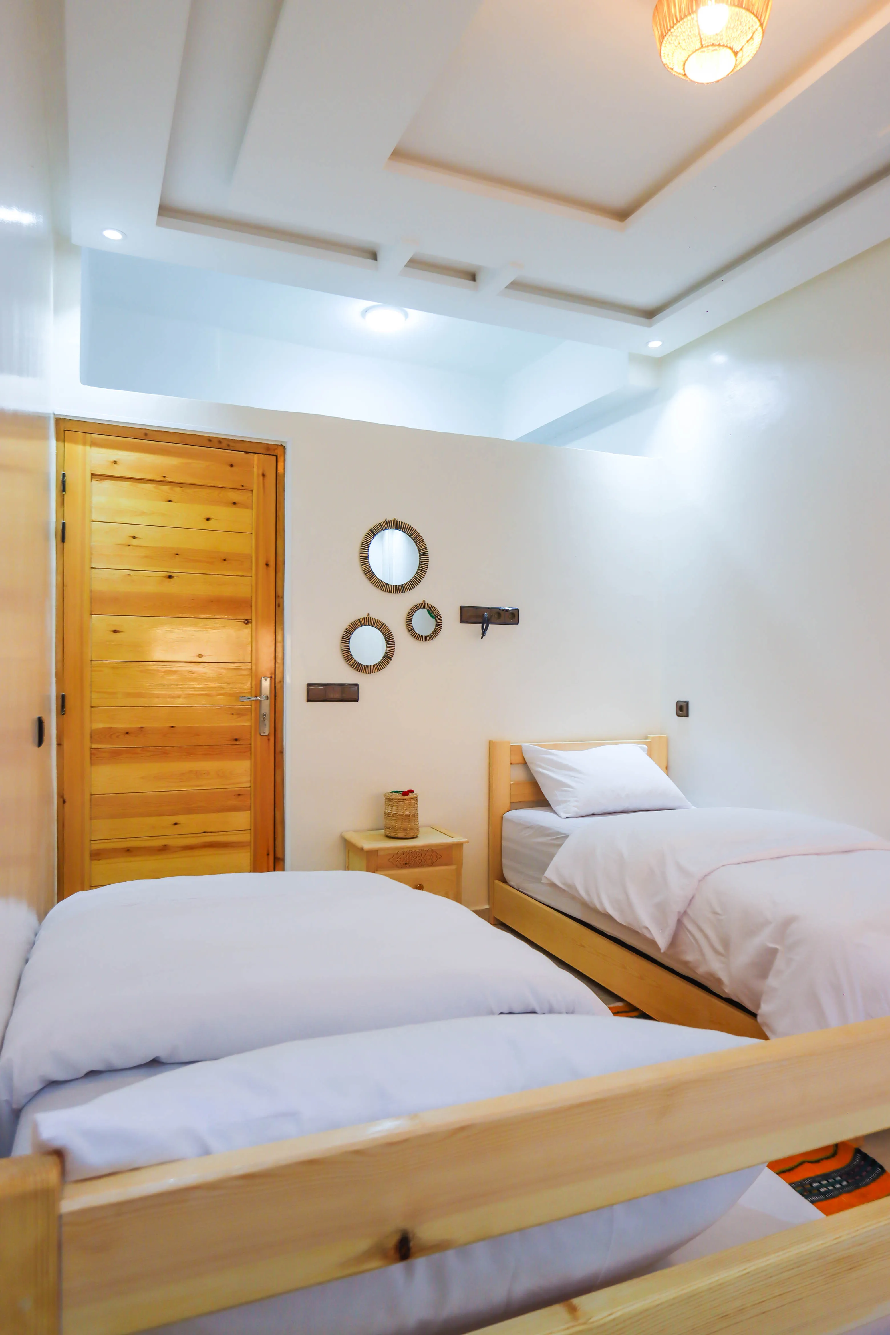 Bright room with two wooden single beds, white bedding, a wooden door, and three round mirrors on the wall.