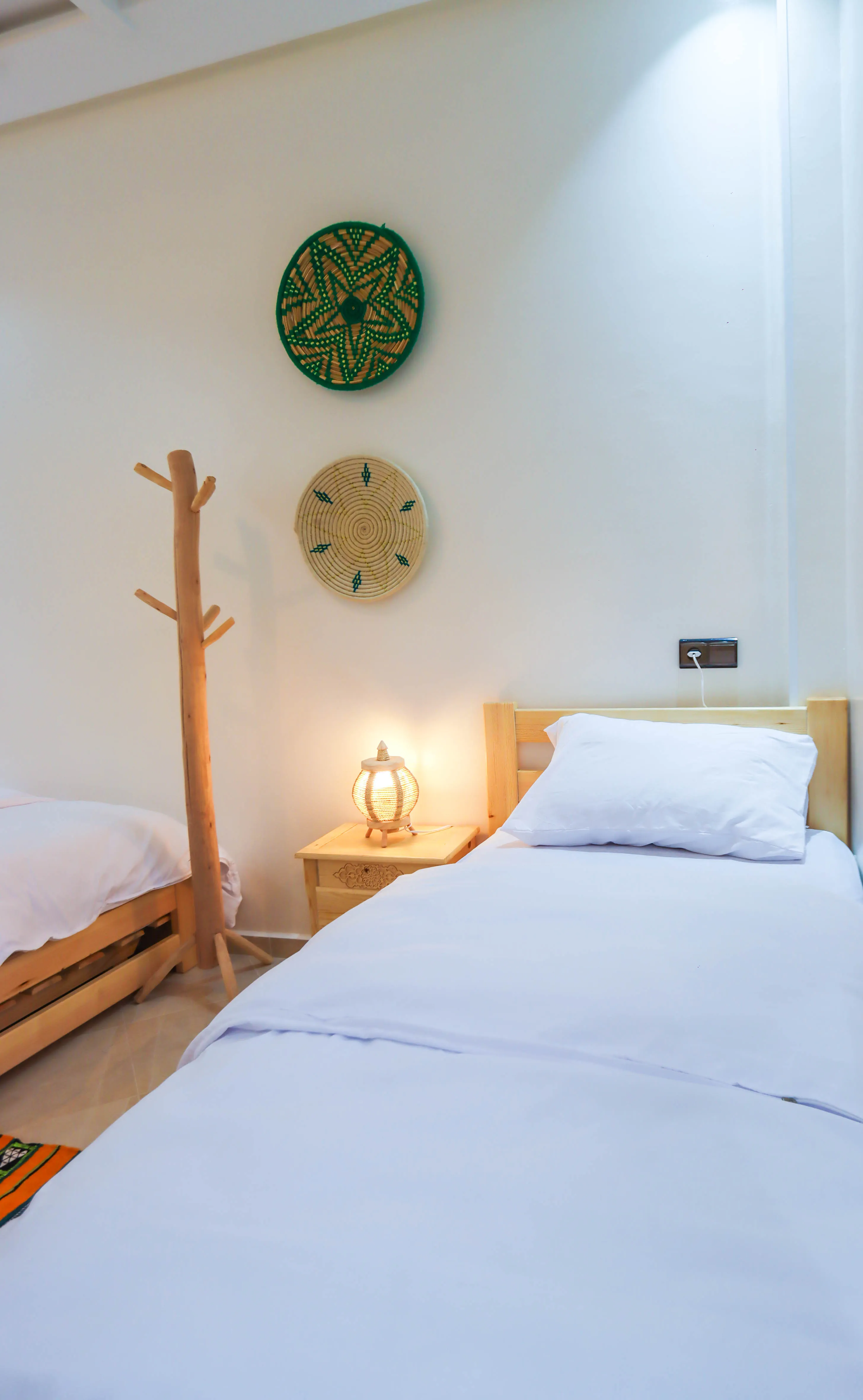 Minimalist bedroom with two wooden beds, white bedding, nightstand with lamp, and decorative woven wall hangings.