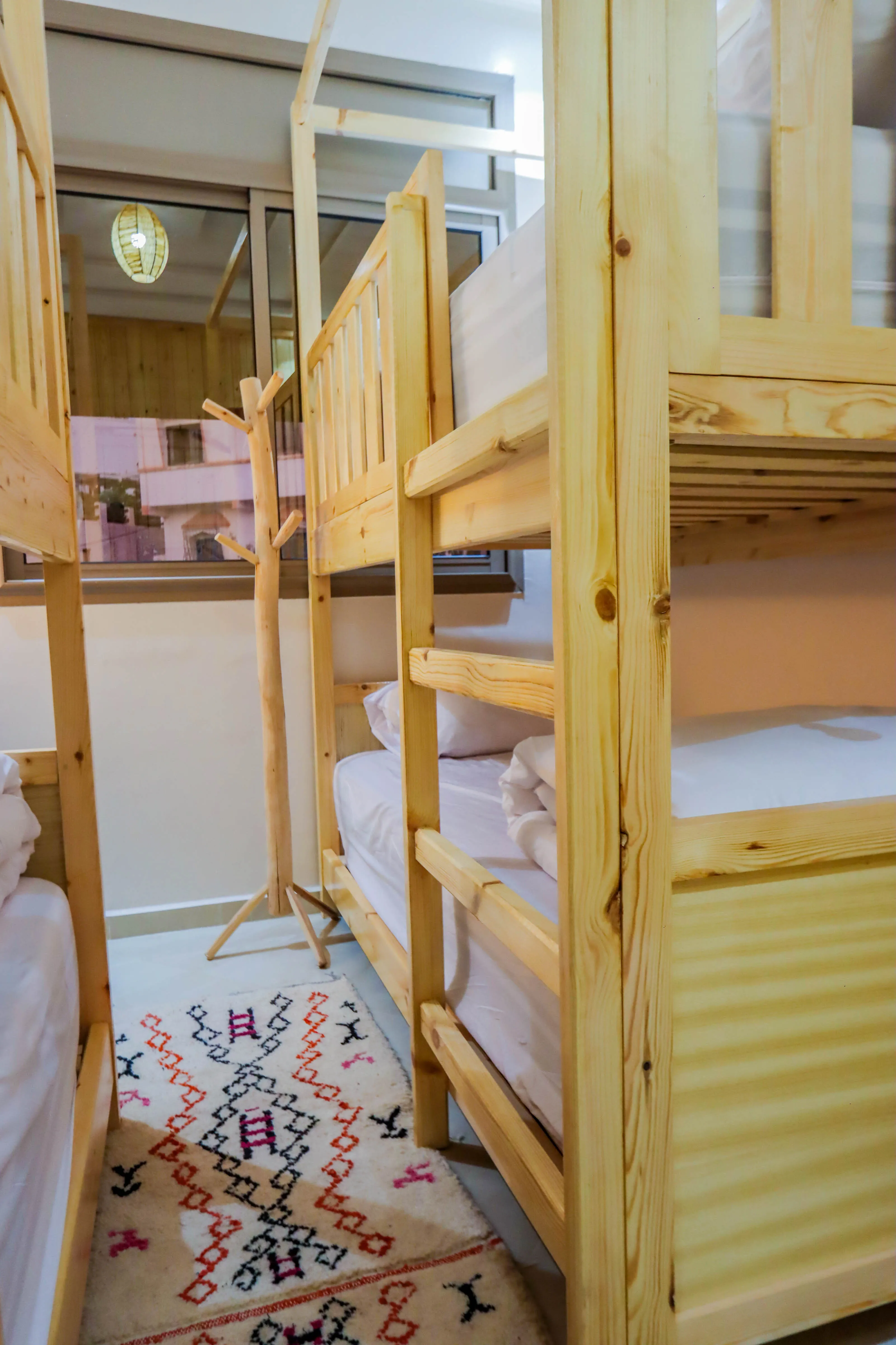 Wooden bunk beds with white bedding in a small room featuring a patterned rug and a wooden coat rack.