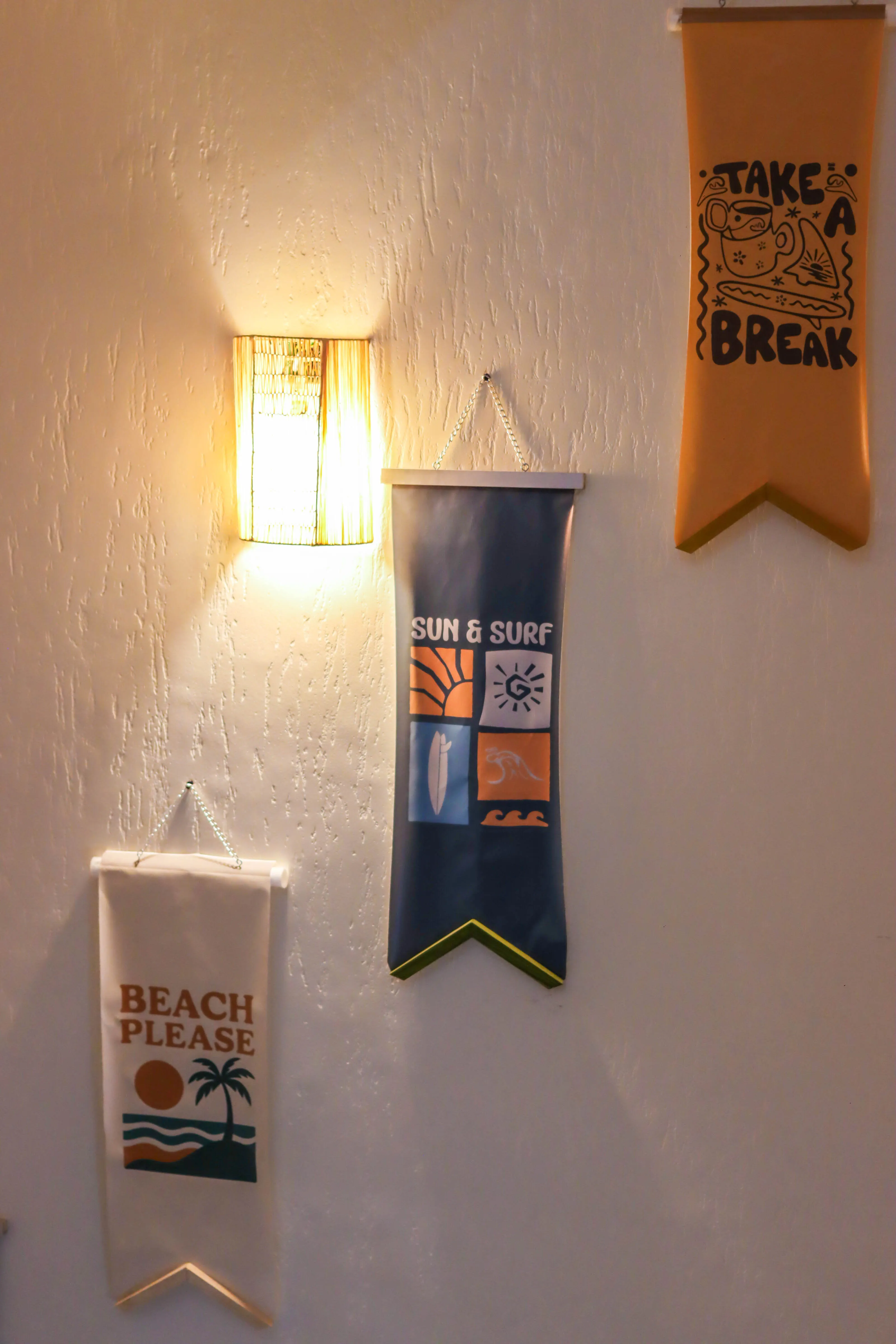 Three hanging decorative banners on a textured wall beside a lit wall sconce light.