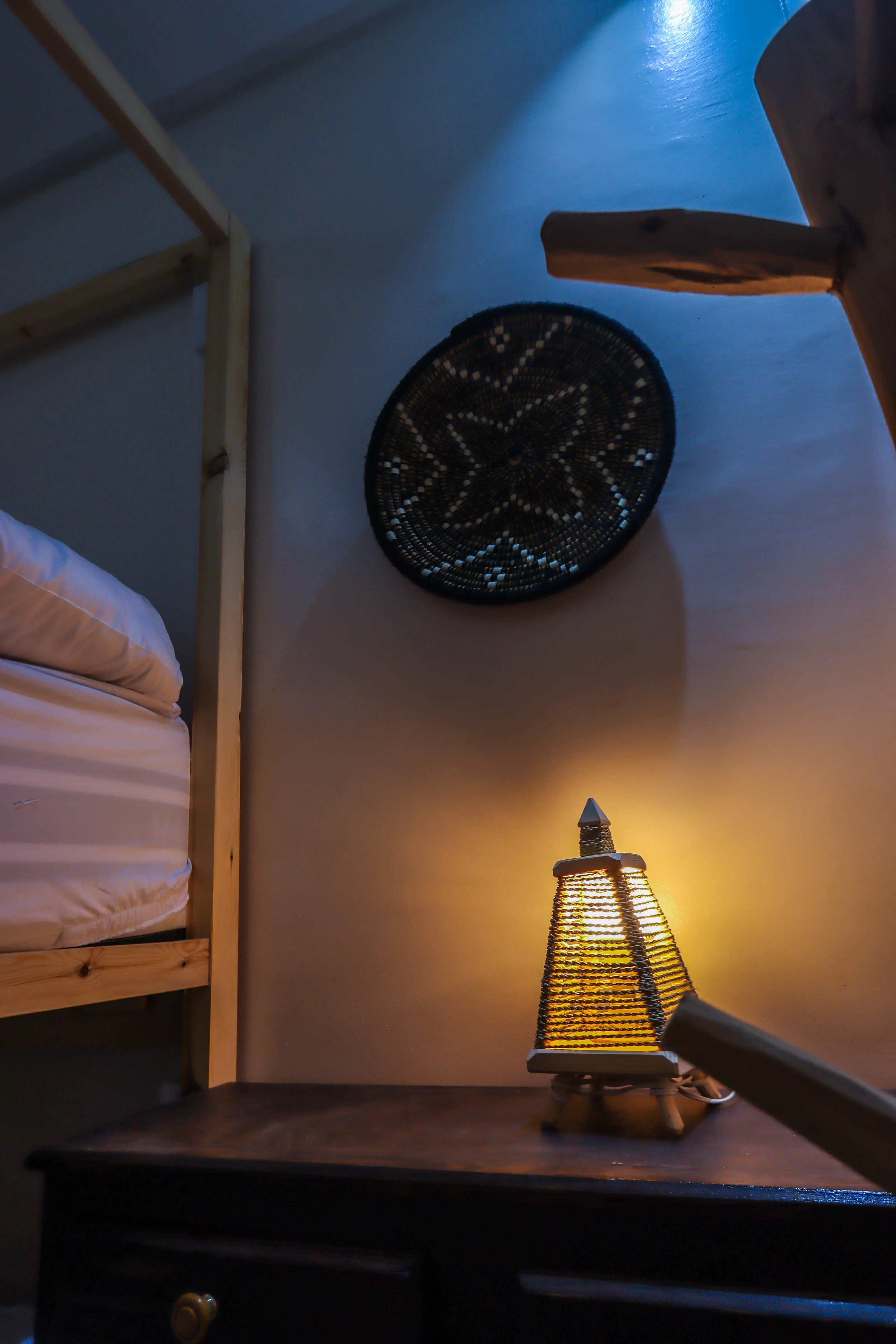 Wooden nightstand with a decorative woven lamp turned on beside a bed with a wooden frame and wall decor above.