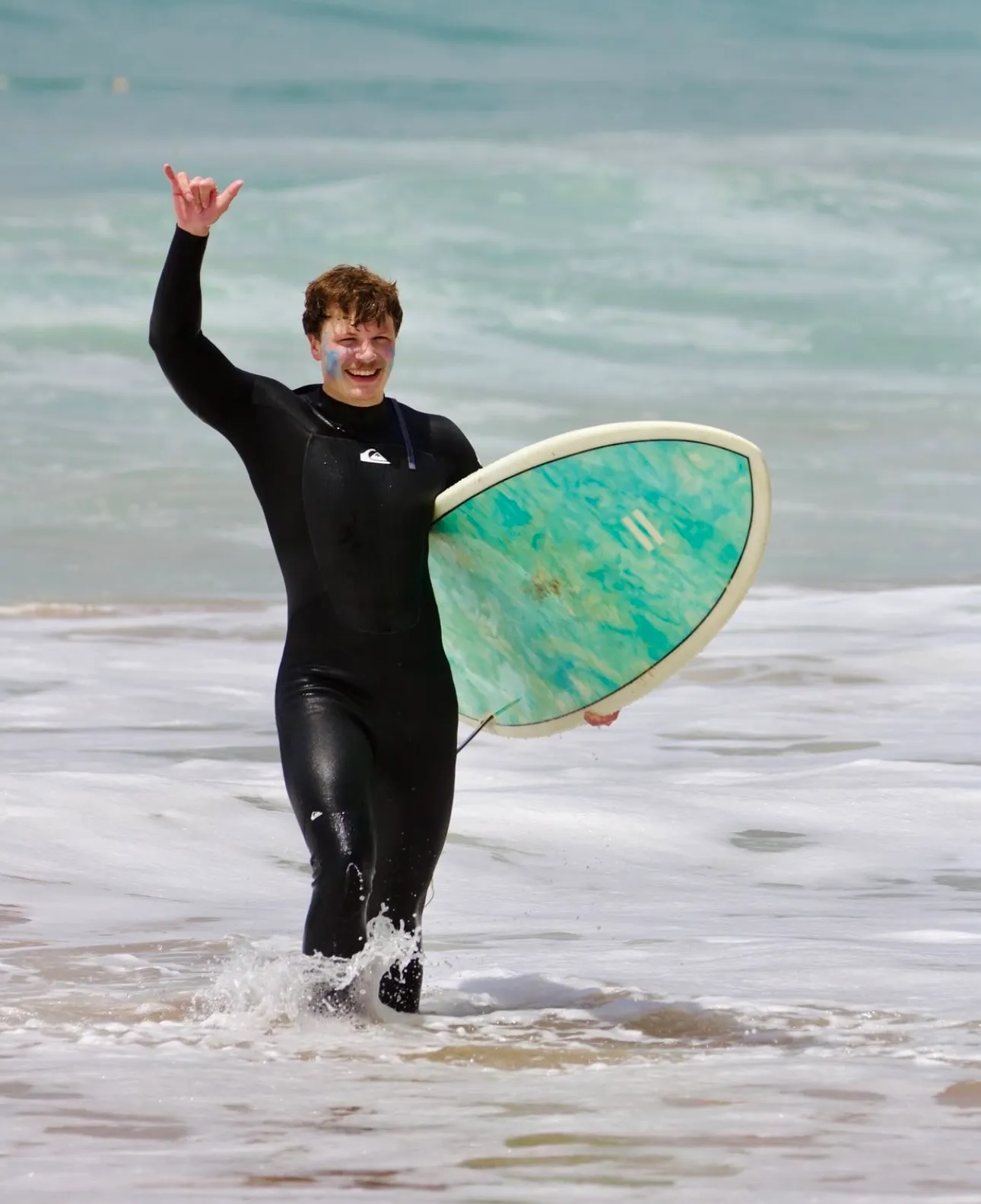 Surfer in black wetsuit holding surfboard and making shaka hand sign walking out of ocean waves.