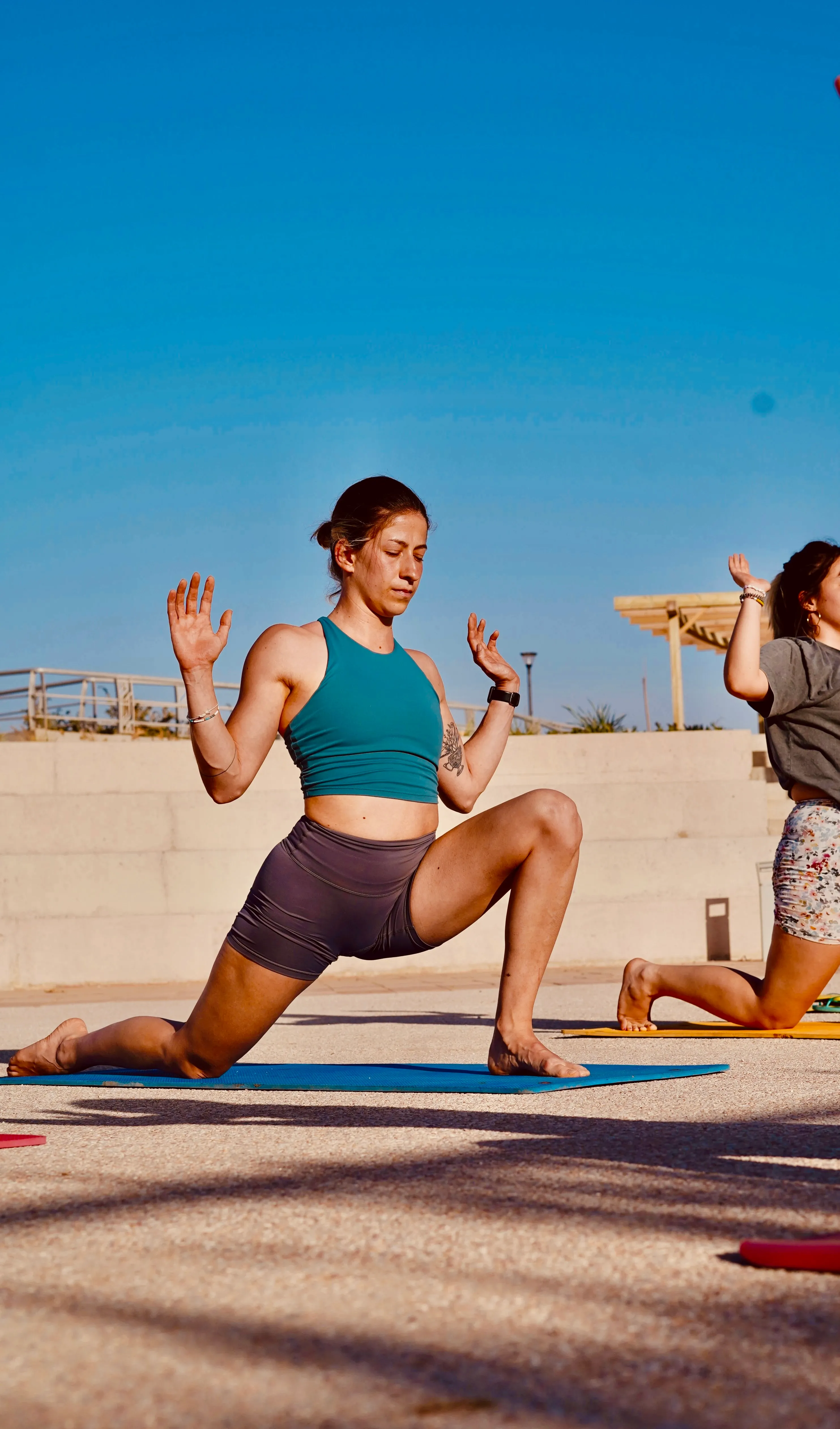 Woman in teal top and dark shorts doing a low lunge yoga pose on blue mat outdoors under clear sky.