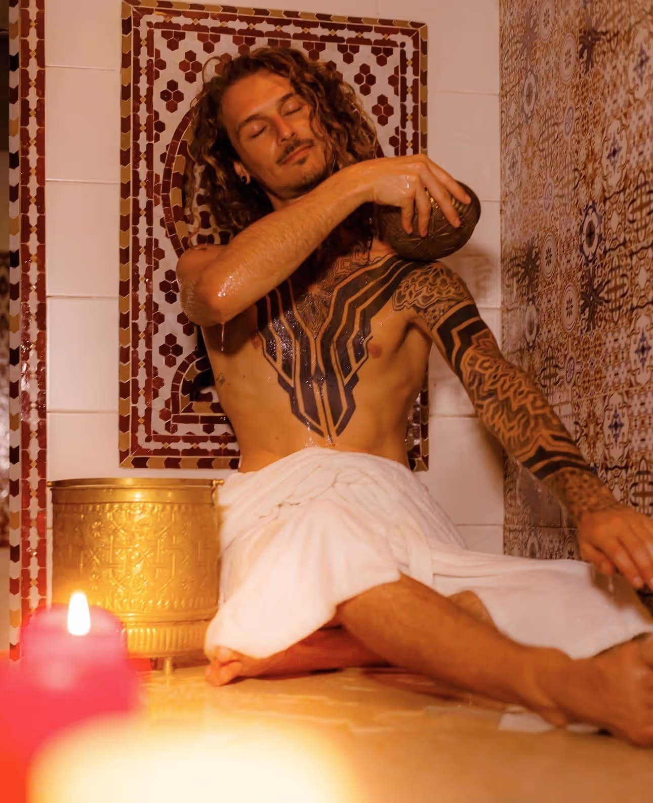 Man with tattoos and a towel pouring water on his shoulder in a decorated bath setting.