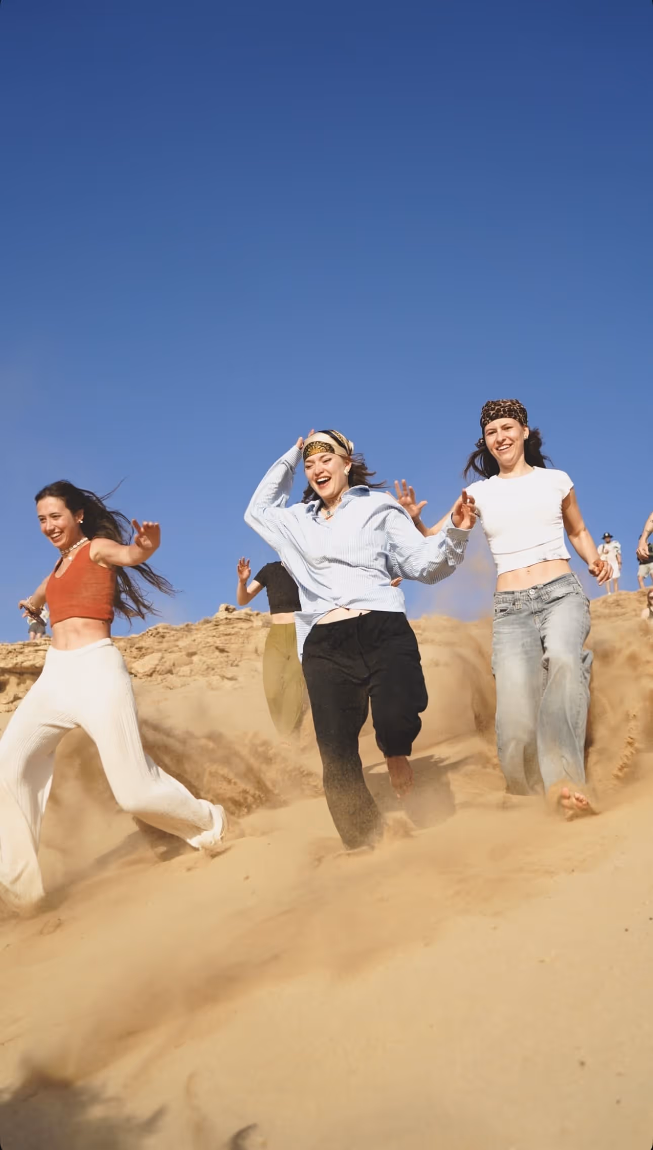 Three young women smiling as they run down a sandy hill under a clear blue sky.