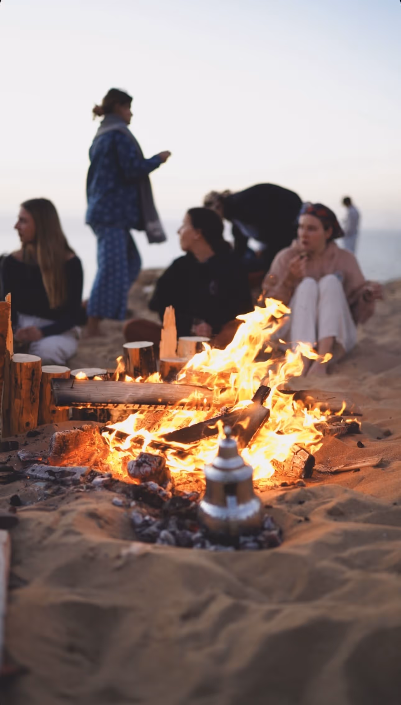 People gathered around a large campfire on sand at dusk with a metal pot near the fire.