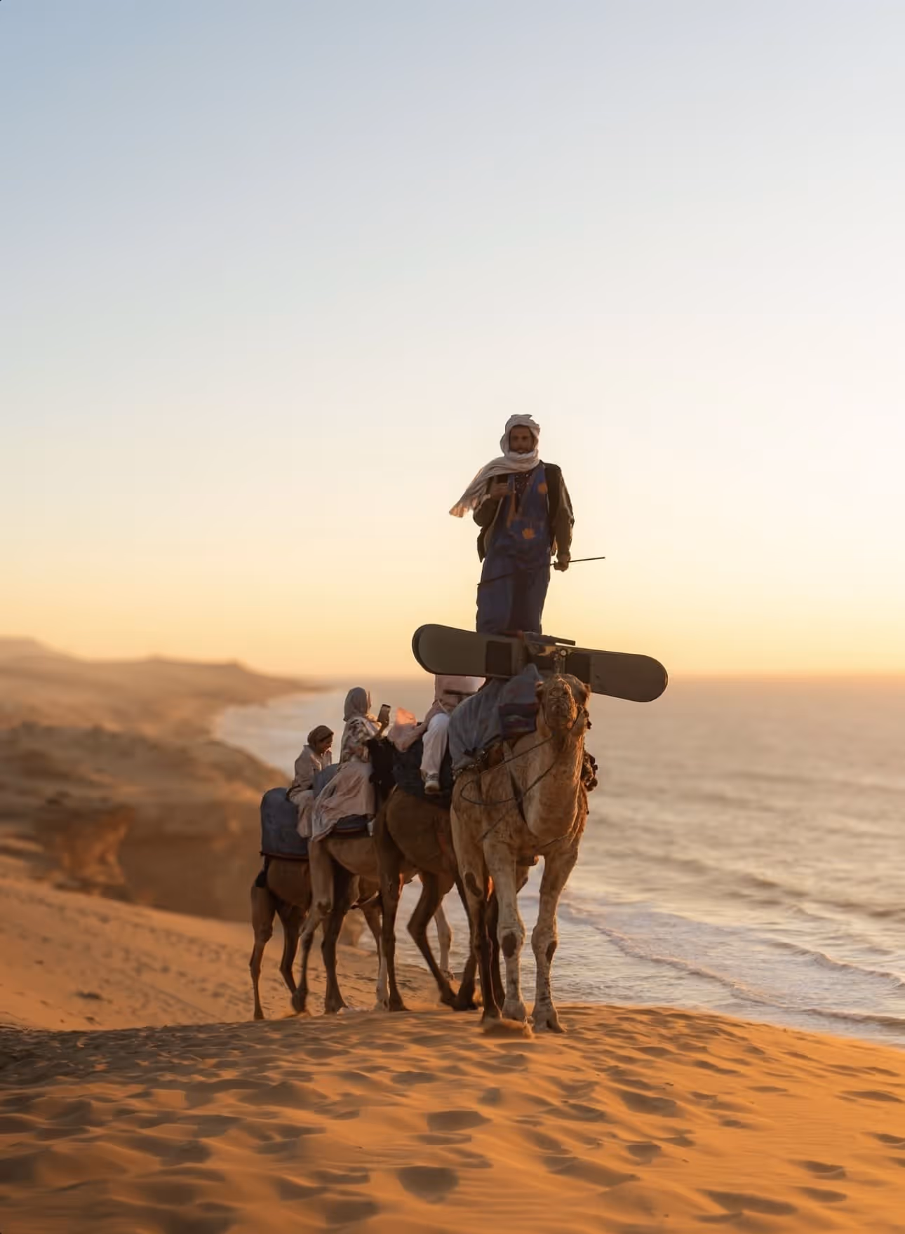 Group of people riding camels on sand dunes near the ocean at sunset.