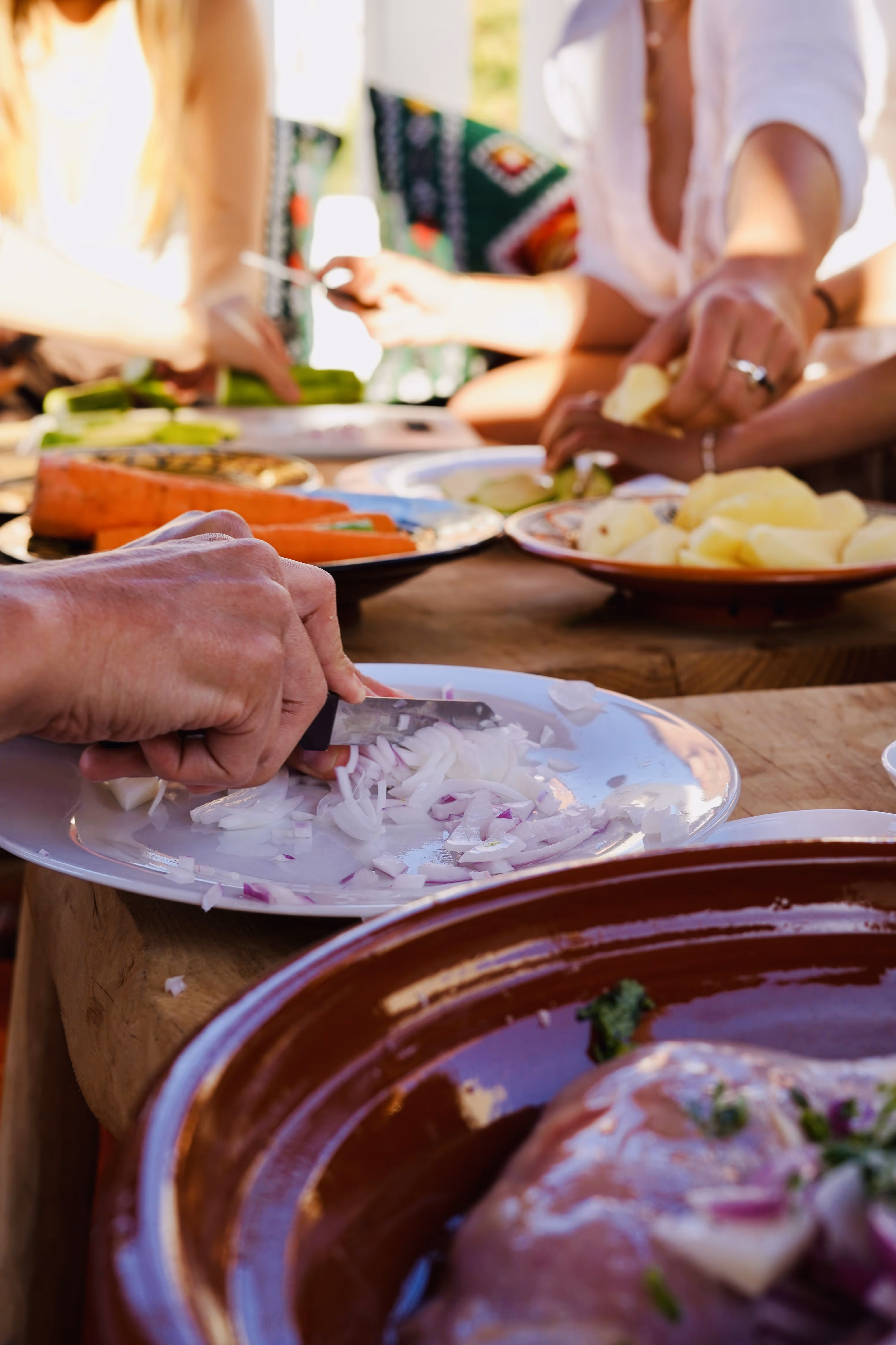Hands chopping onions on a white plate with vegetables and people preparing food in the background.